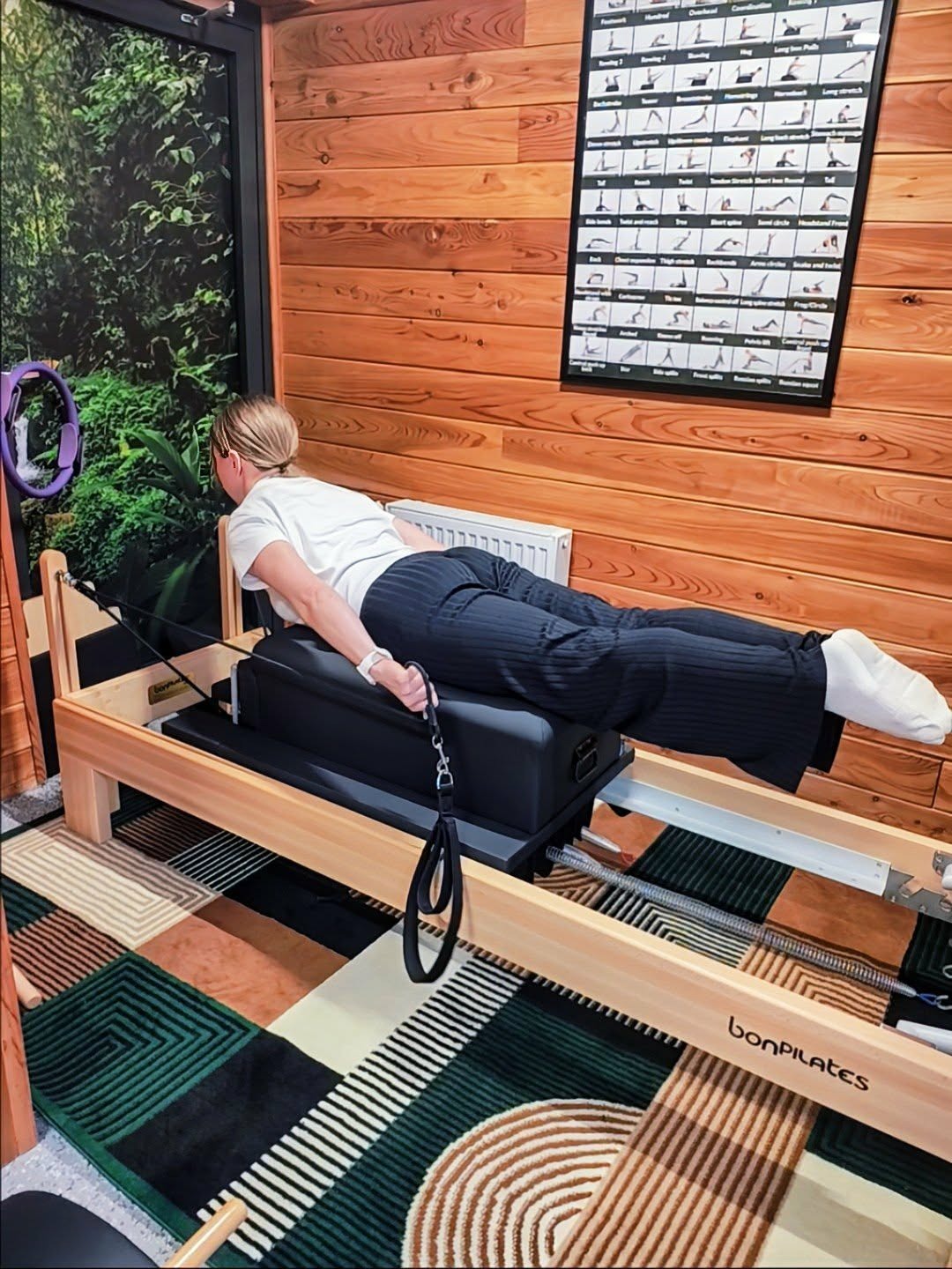 Kobieta ćwiczy na reformerze BonPilates w studiu z drewnianymi ścianami i plakatem z ćwiczeniami. Widoczny dywan w geometryczne wzory i element dekoracyjny z roślinami.