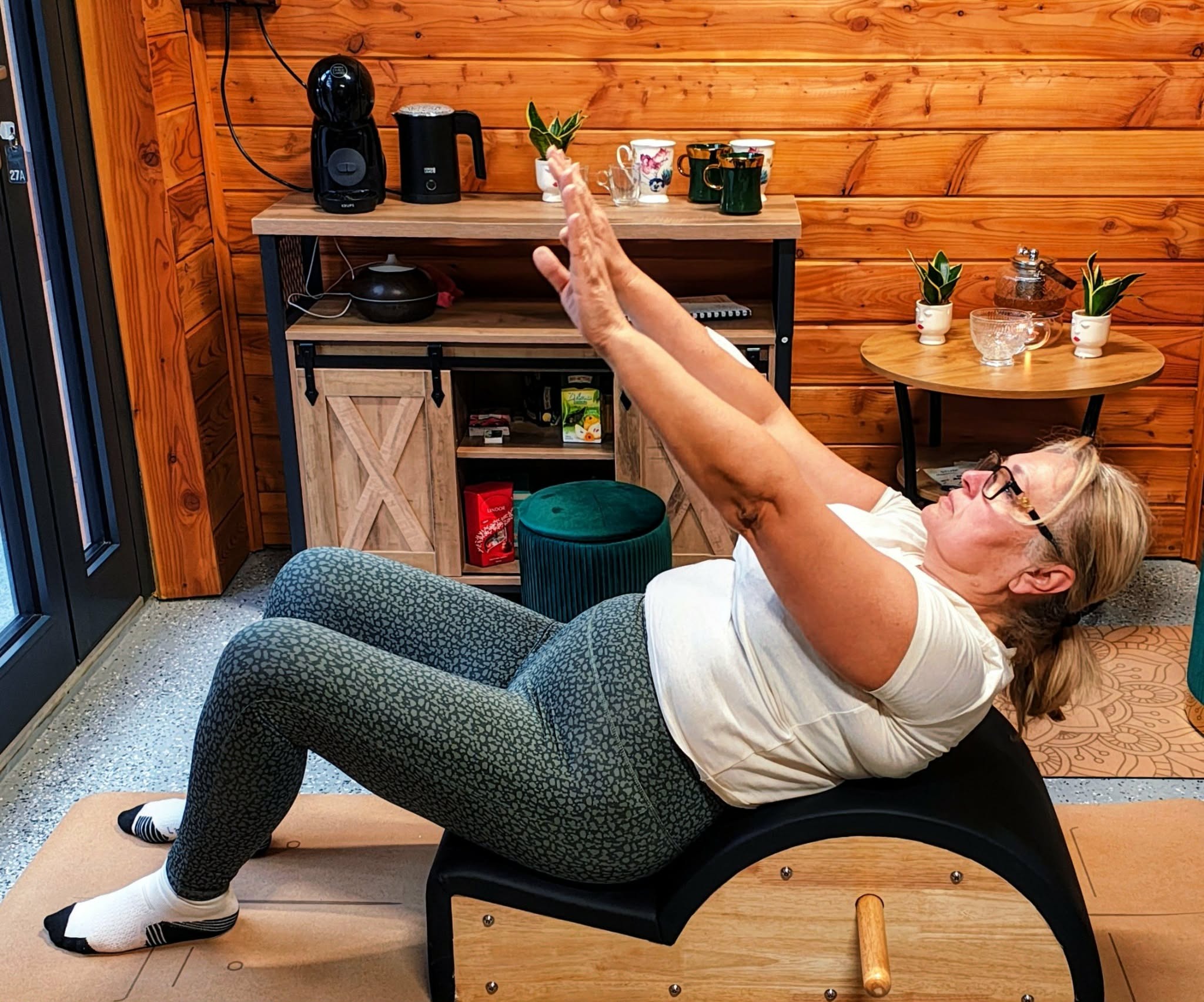 Kobieta ćwiczy na reformerze pilates w studio z drewnianymi ścianami. Widoczne akcesoria do ćwiczeń i napoje. Skupienie na prawidłowej postawie podczas ćwiczeń.