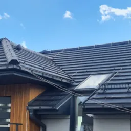 Nowoczesny dom z ciemnoszarą dachówką ceramiczną, widoczne okna dachowe, komin obłożony dachówką i systemy odprowadzania wody, na tle błękitnego nieba z chmurami.