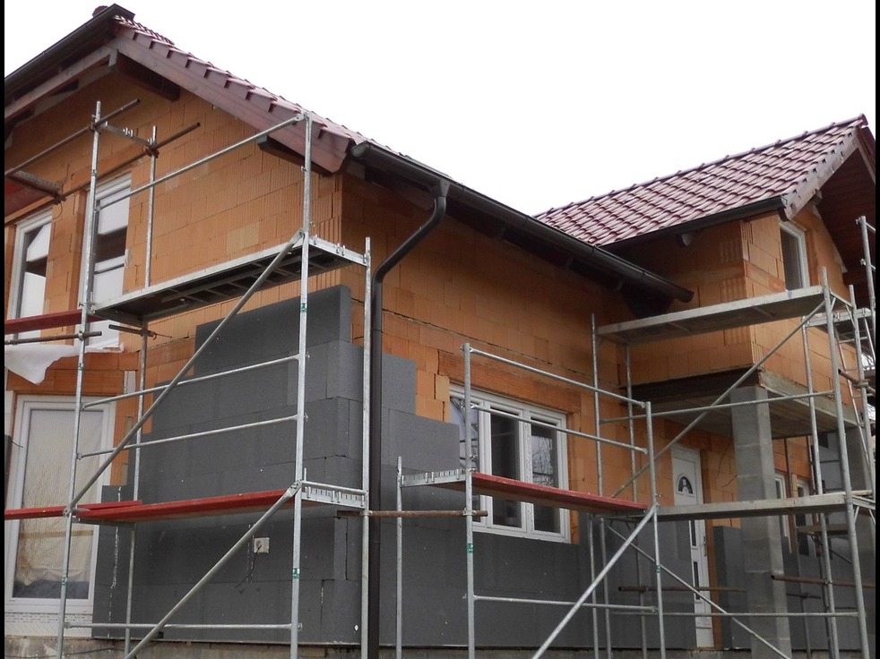 Dom w trakcie ocieplania szarym styropianem, widoczne rusztowania i fragment nieocieplonej ściany z cegły ceramicznej.