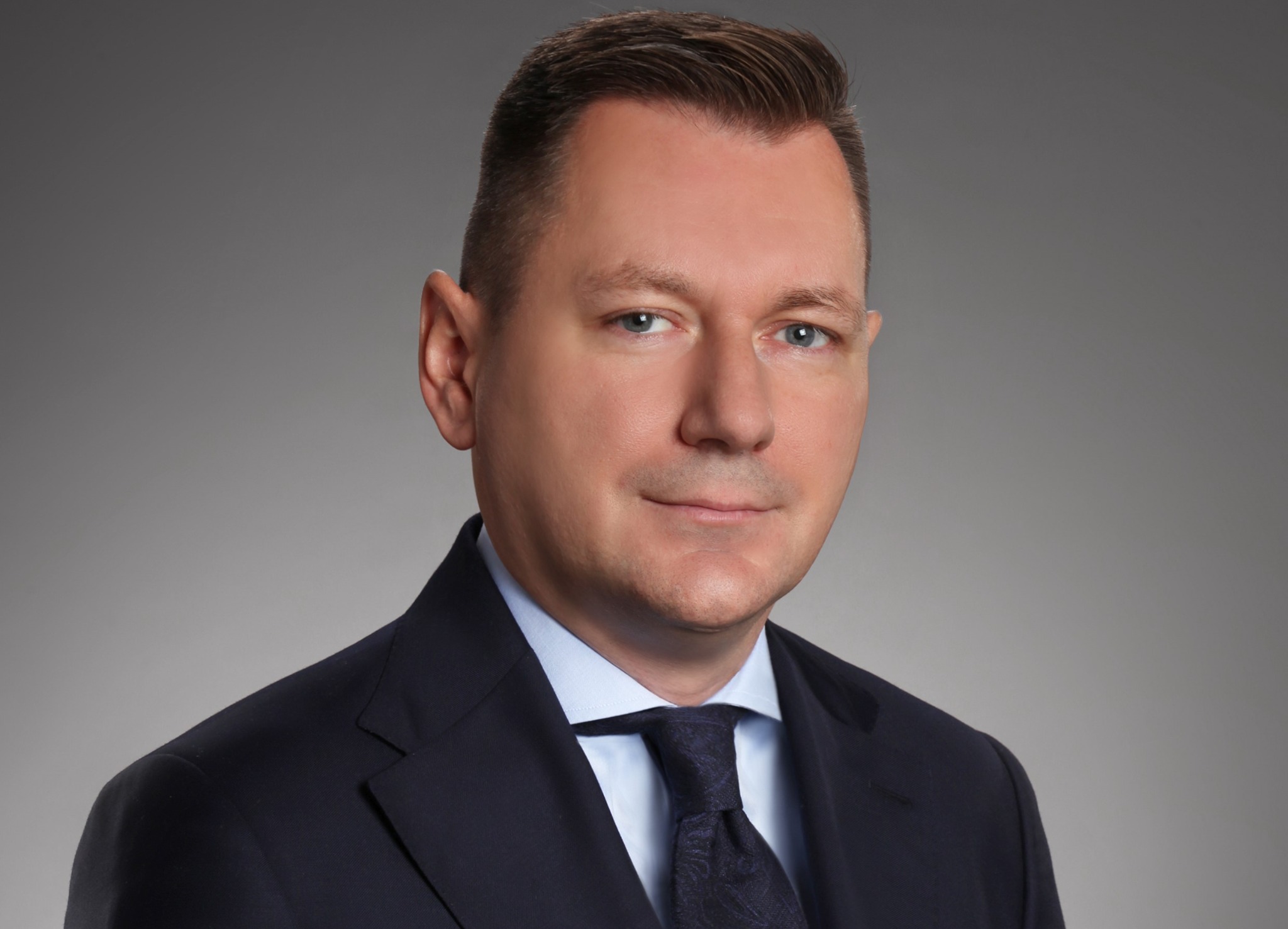 Portret mężczyzny w garniturze i krawacie na szarym tle, z lekkim uśmiechem i niebieskimi oczami.