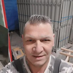 Portret pracownika z siwymi włosami na tle metalowych ram okiennych związanych niebieską taśmą w magazynie.