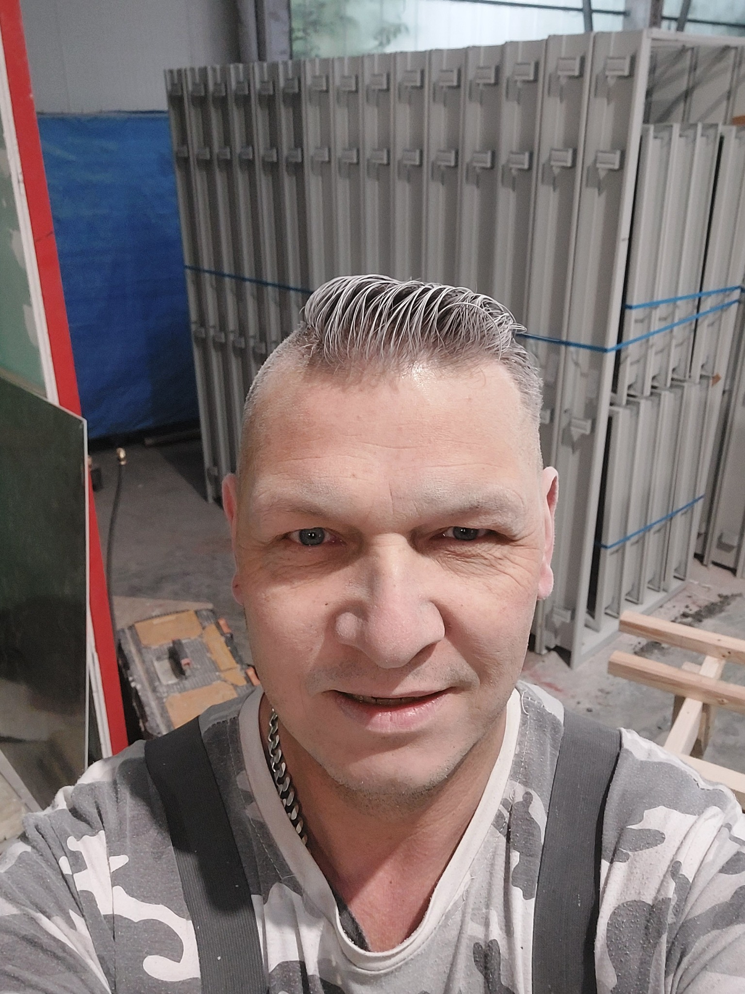 Portret pracownika z siwymi włosami na tle metalowych ram okiennych związanych niebieską taśmą w magazynie.