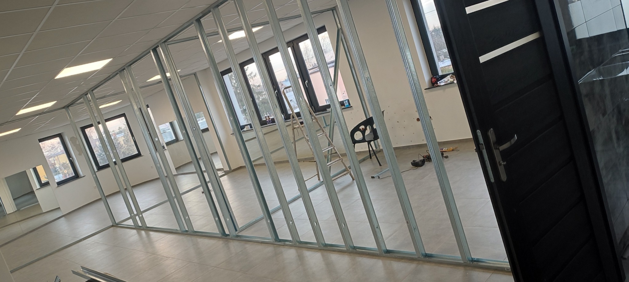 Stelaż z profili stalowych do ścianki działowej w jasnym pomieszczeniu biurowym, widoczna drabina, czarne drzwi i narzędzia na podłodze.