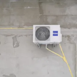 Biała jednostka zewnętrzna klimatyzatora Haier zamontowana na surowej, szarej ścianie budynku w trakcie budowy, z widocznymi przewodami zabezpieczonymi żółtą pianką montażową.