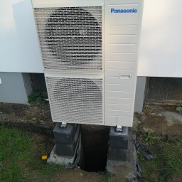 Thermro Dawid Mrozowski - Zewnętrzna jednostka pompy ciepła Panasonic, zamontowana na betonowych blokach fundamentowych, z widocznymi wykopami fundamentowymi i żółtą linką wyznaczającą teren.