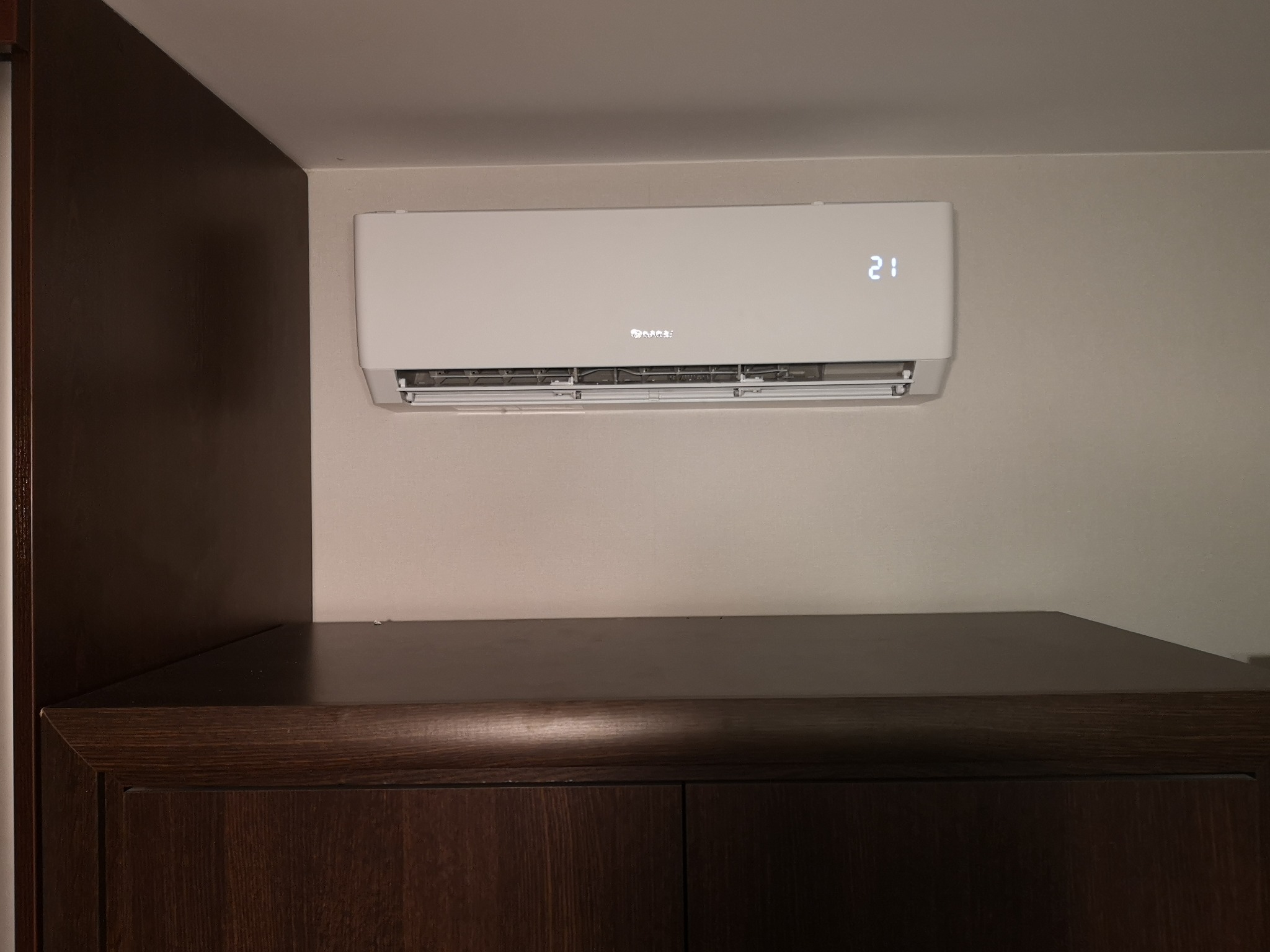 Biały klimatyzator ścienny z wyświetlaczem temperatury 21 stopni Celsjusza, zamontowany nad ciemnobrązową szafką.