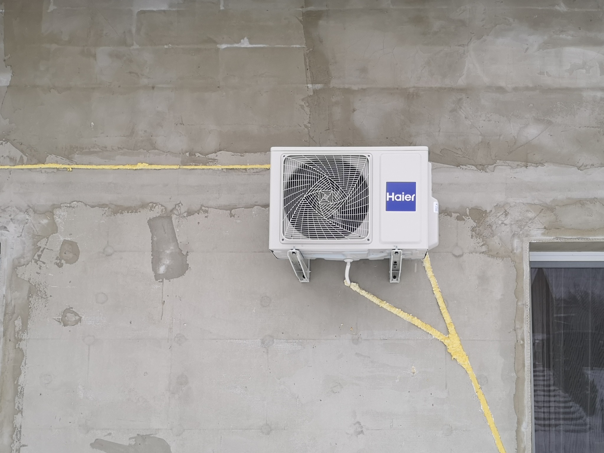 Biała jednostka zewnętrzna klimatyzatora Haier zamontowana na surowej, szarej ścianie budynku w trakcie budowy, z widocznymi przewodami zabezpieczonymi żółtą pianką montażową.