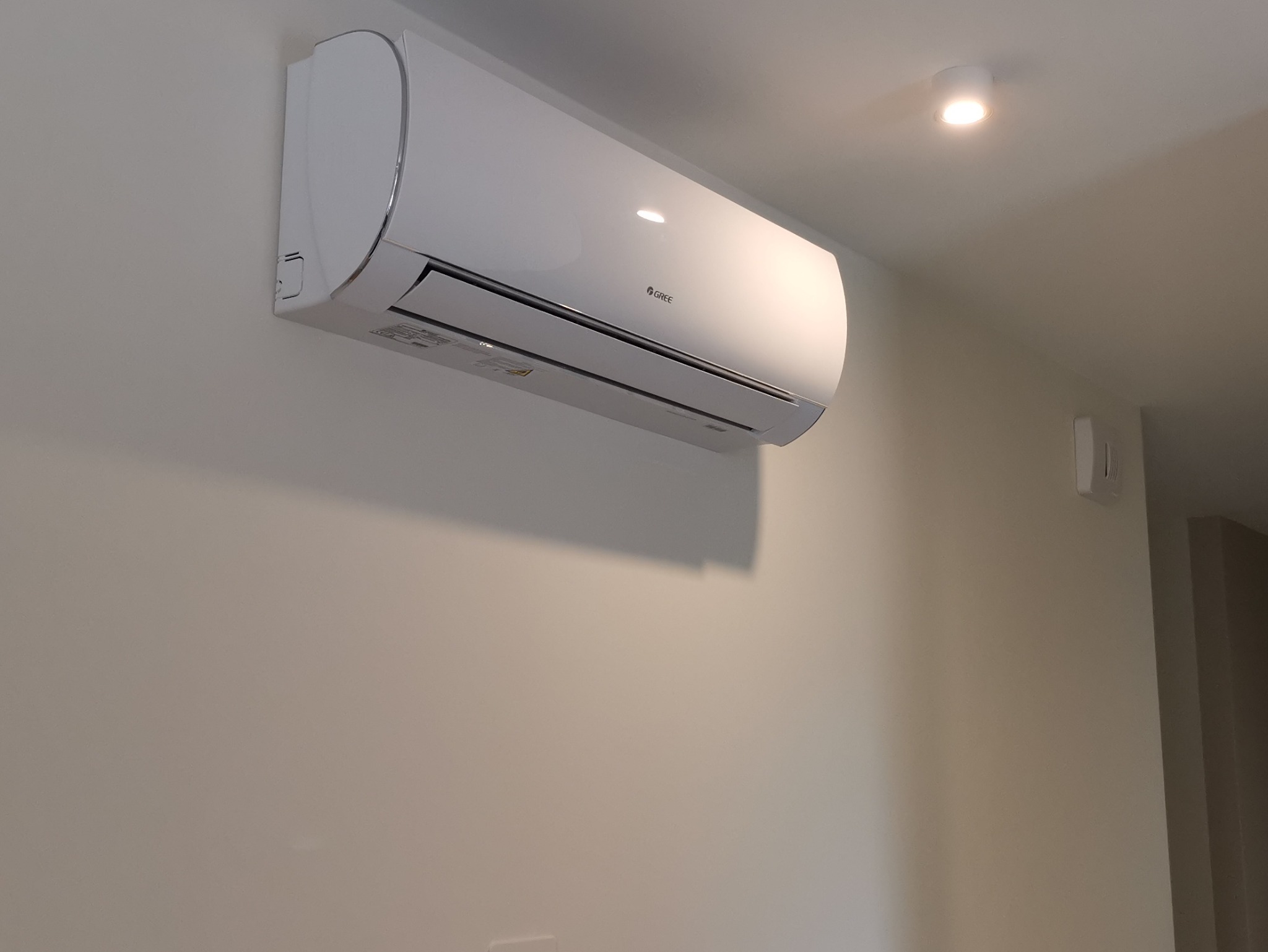 Biała klimatyzacja marki Gree zamontowana na jasnej ścianie w pomieszczeniu. Widoczny fragment sufitu z oświetleniem punktowym i czujnik.