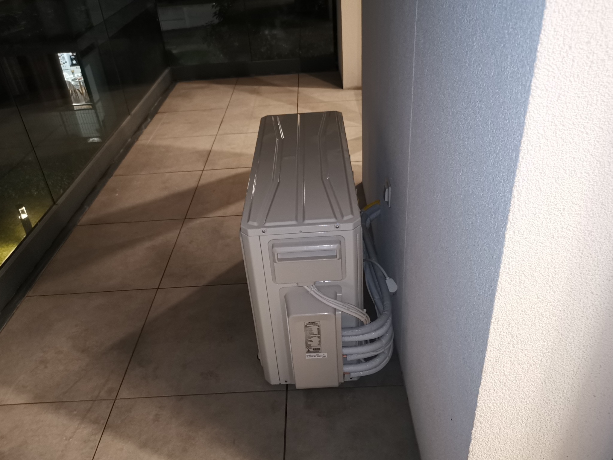 Zewnętrzna jednostka klimatyzacji zamontowana na balkonie, widoczne przewody i fragment ściany budynku. Ujęcie z boku, w tle płytki balkonowe.