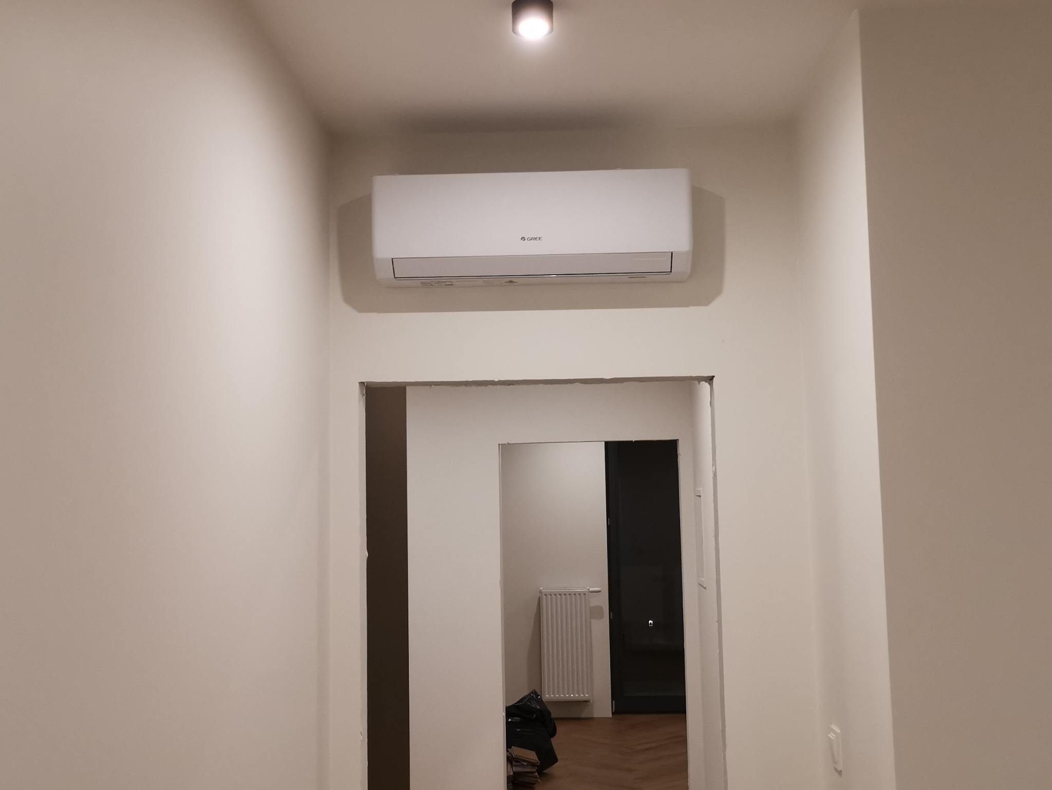 Biała klimatyzacja marki Gree zamontowana nad przejściem w jasnym wnętrzu. Widoczny fragment sufitu z lampą. W tle widoczne pomieszczenie z grzejnikiem.