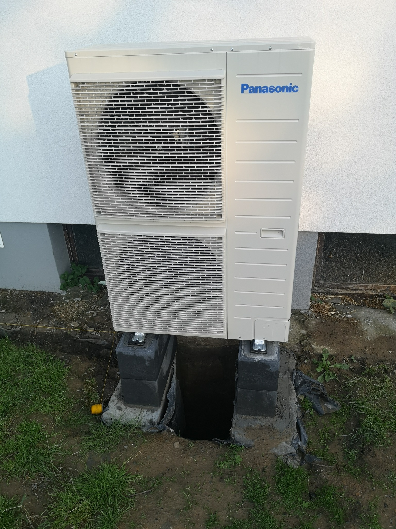 Zewnętrzna jednostka pompy ciepła Panasonic, zamontowana na betonowych blokach fundamentowych, z widocznymi wykopami fundamentowymi i żółtą linką wyznaczającą teren.