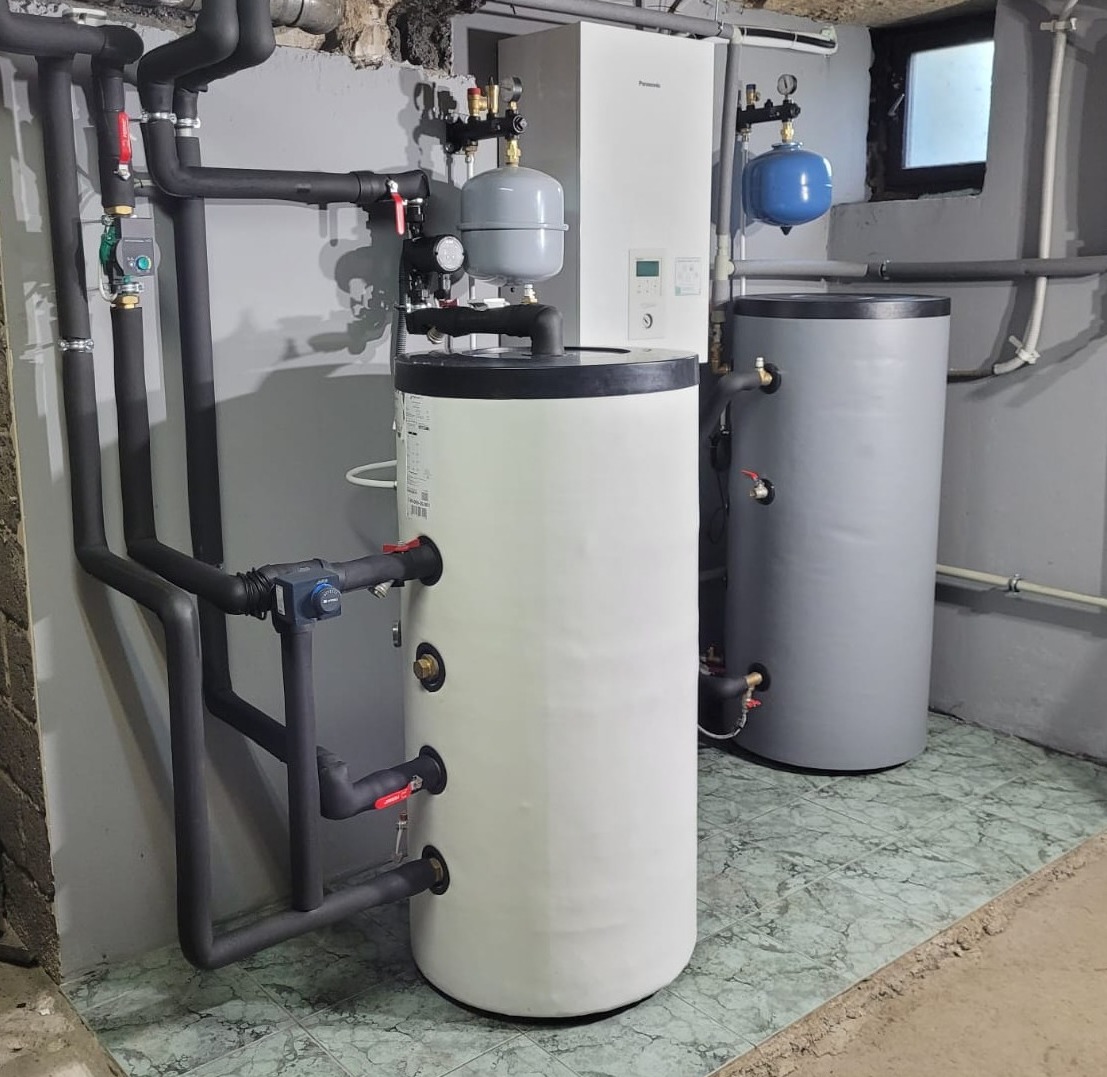 Instalacja centralnego ogrzewania z dwoma zbiornikami akumulacyjnymi w piwnicy, rury w izolacji termicznej, widoczny manometr i zawory.