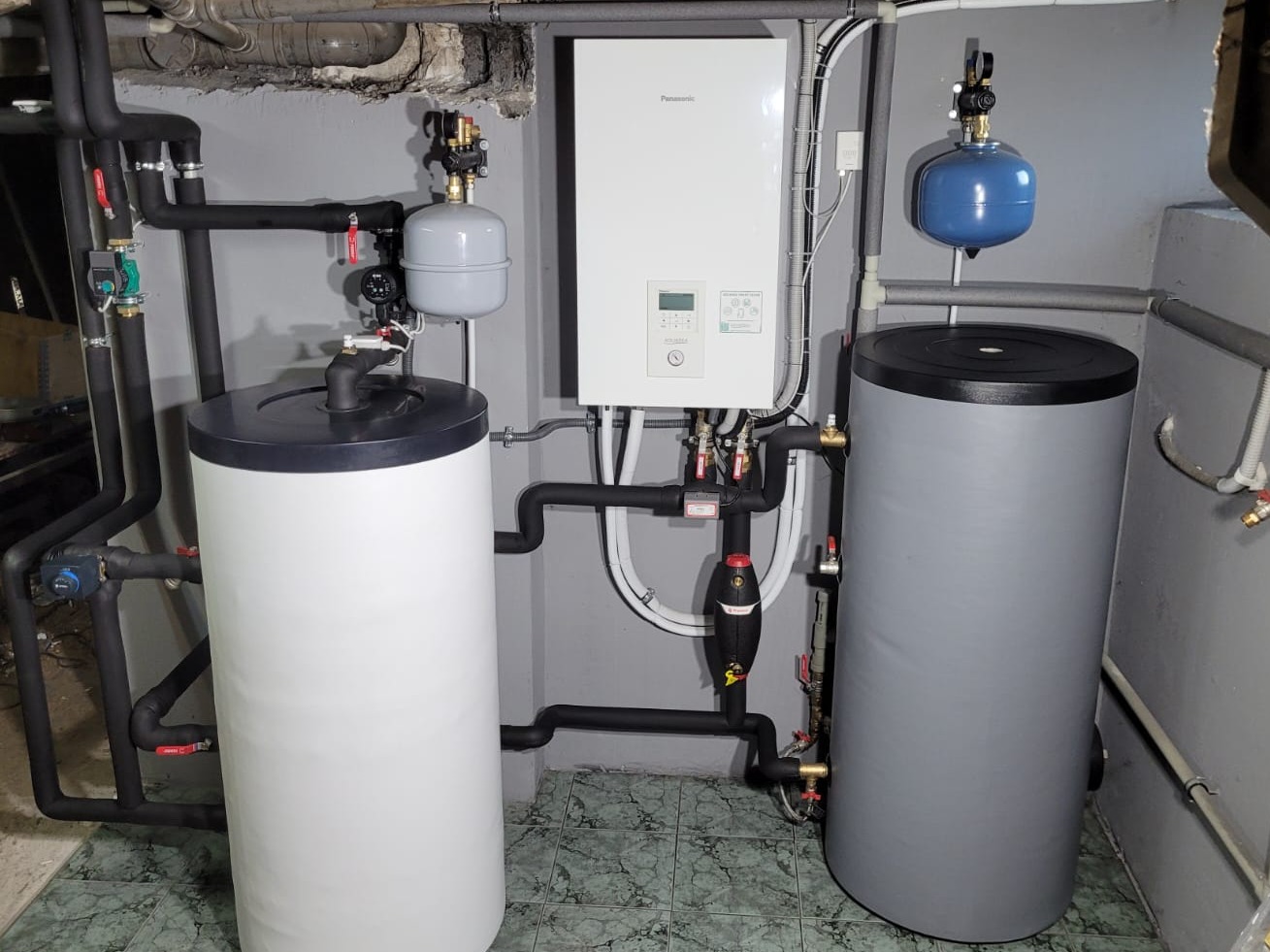Zestaw urządzeń grzewczych: biały zasobnik ciepłej wody, szary zasobnik, biały panel sterowania Panasonic oraz orurowanie w piwnicy z szarą ścianą i zieloną podłogą.