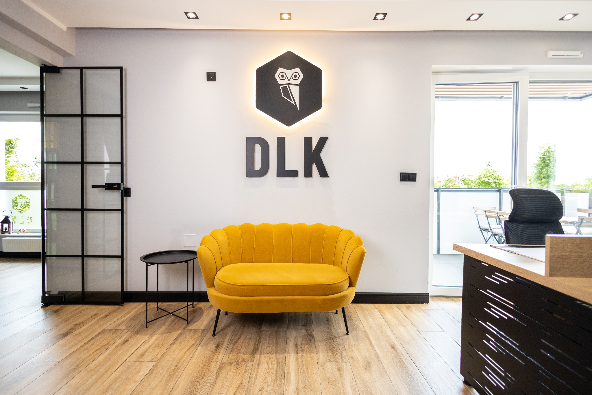 Nowoczesne wnętrze biura z żółtą sofą na drewnianej podłodze, logo firmy DLK na białej ścianie i czarną, przeszkloną ścianką działową.