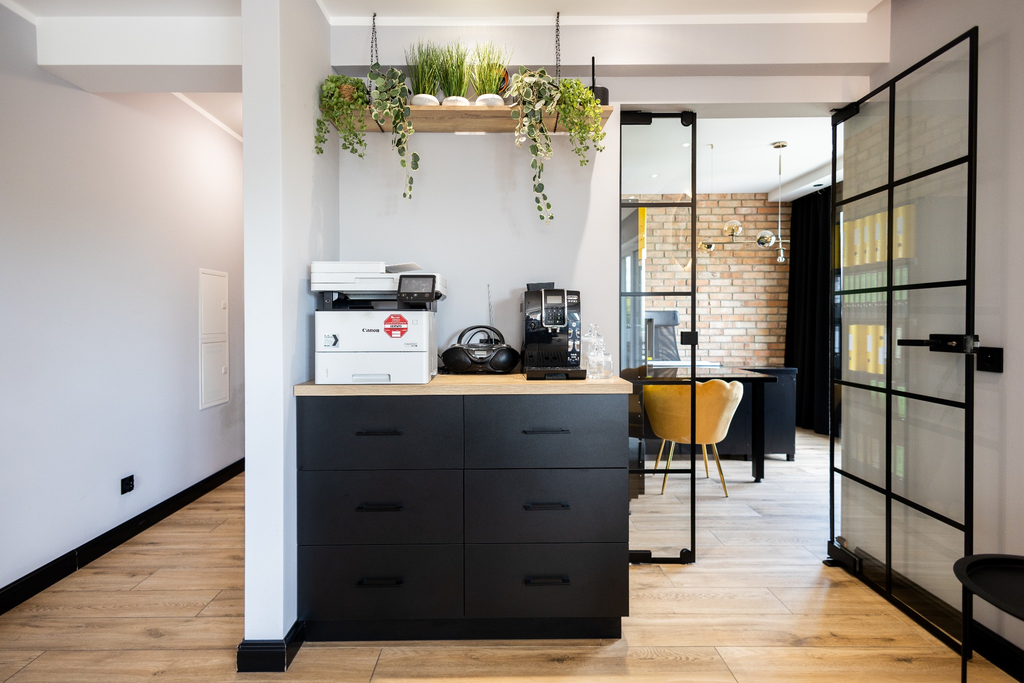 Nowoczesne wykończenie wnętrza biura: czarna komoda z blatem z drewna, sprzęt biurowy, rośliny wiszące, przeszklone drzwi, widok na biurko i ceglaną ścianę w tle.