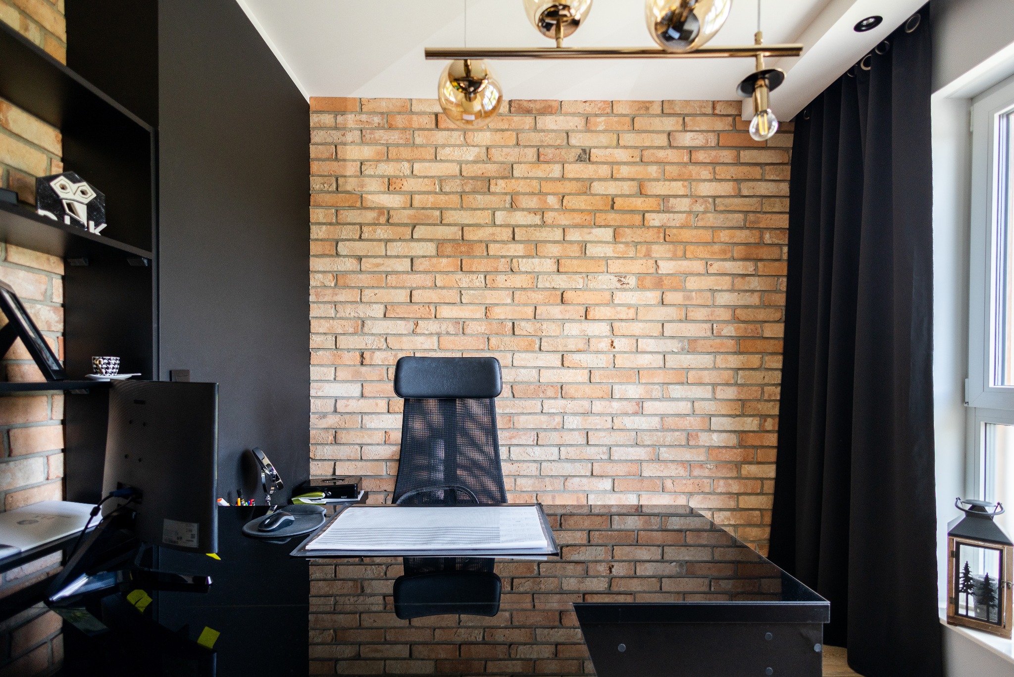 Biuro z ceglaną ścianą, czarnym biurkiem i krzesłem. Na półkach dekoracje, nowoczesna lampa sufitowa i okno z czarnymi zasłonami tworzą stylowe wnętrze.