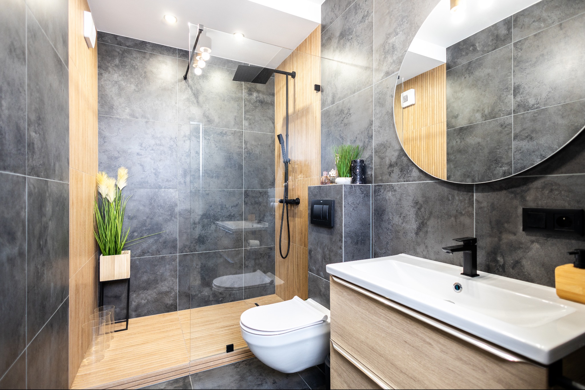 Nowoczesna łazienka z kabiną typu walk-in, drewnianym akcentem, szarą płytką i okrągłym lustrem. Minimalistyczny design i naturalne materiały.