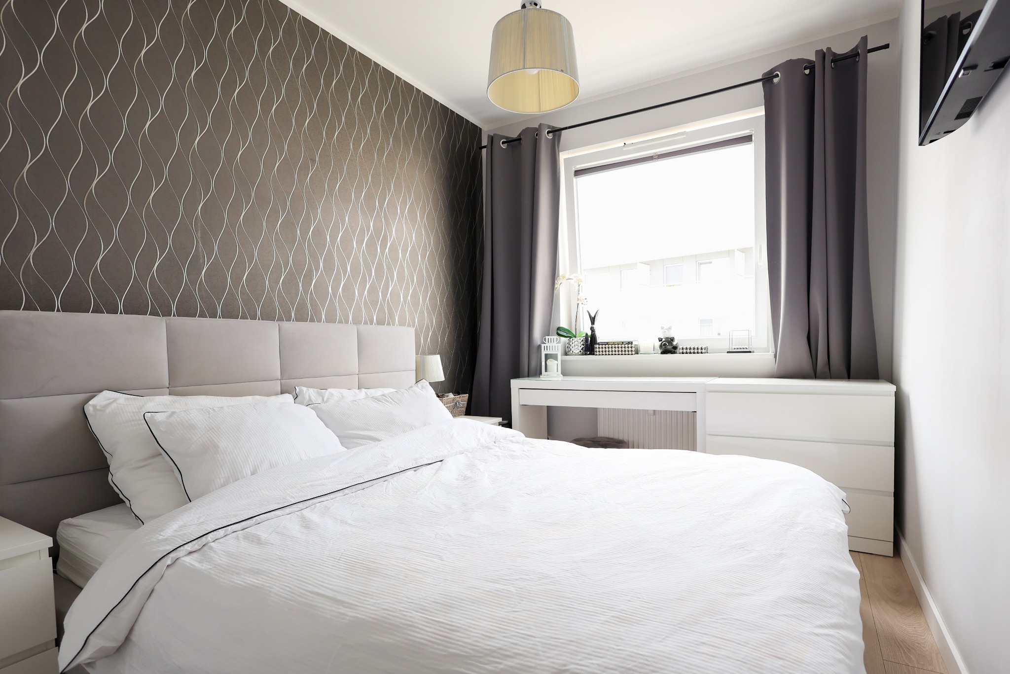Nowoczesna sypialnia z łóżkiem, tapetą w fale i białym biurkiem pod oknem. Szare zasłony, lampa sufitowa i minimalistyczne dodatki tworzą spójny, elegancki wystrój.