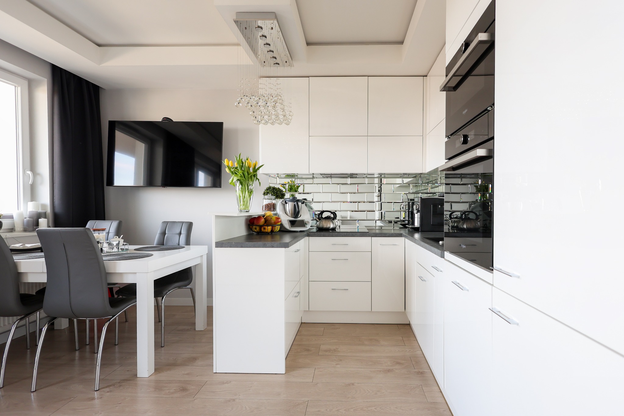 Nowoczesna, jasna kuchnia z białymi meblami i lustrzanymi płytkami, widok z jadalni z białym stołem i szarymi krzesłami. Żółte tulipany w wazonie dodają koloru.