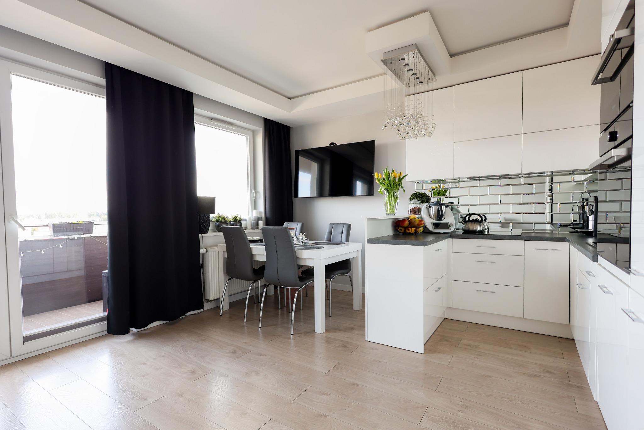 Jasna kuchnia z białymi meblami, lustrzanymi płytkami i drewnianą podłogą. Stół z krzesłami przy oknie z czarnymi zasłonami. Nowoczesne oświetlenie.