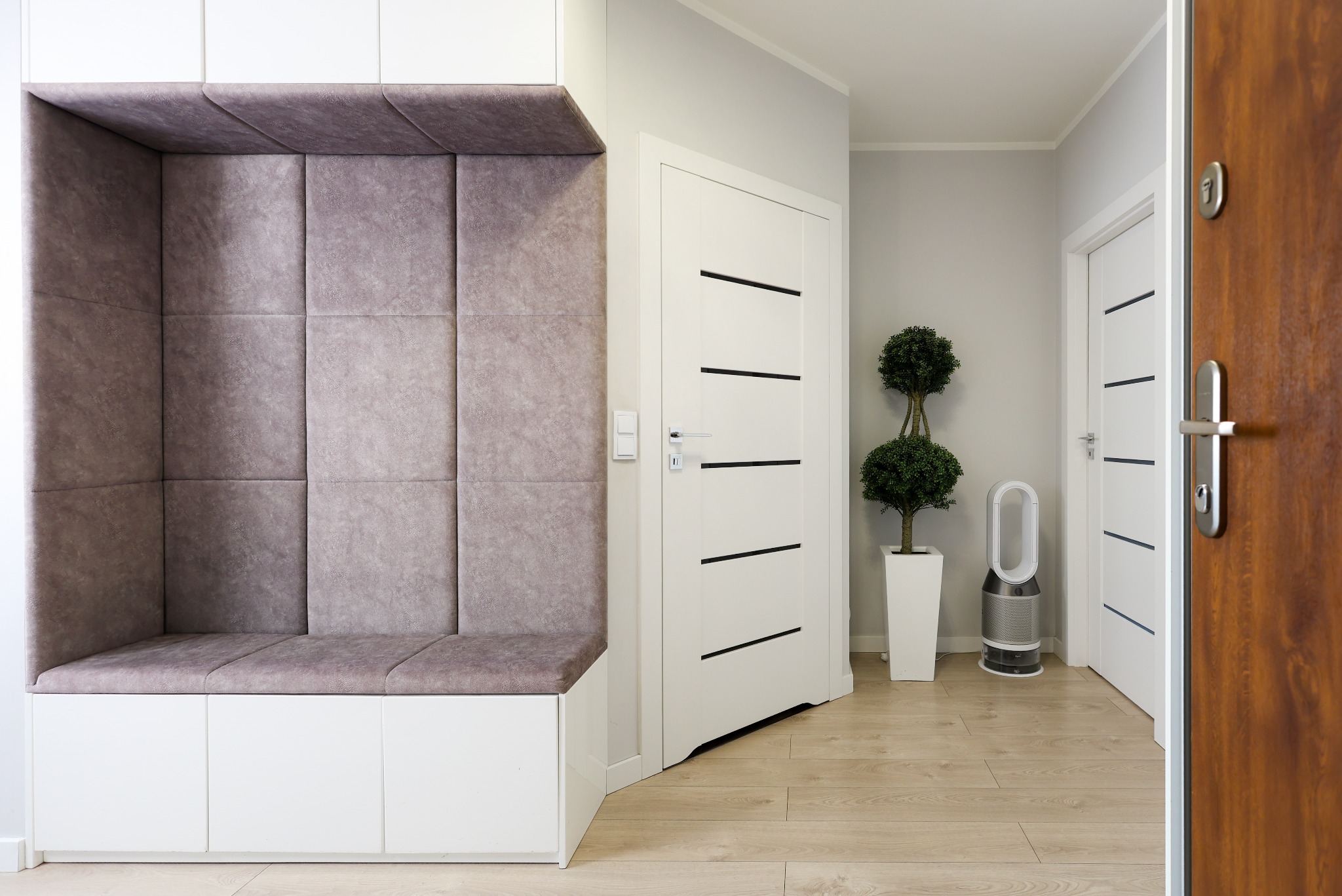 Nowoczesny przedpokój z siedziskiem obitym panelami tapicerowanymi, białe drzwi z czarnymi akcentami i drewniana podłoga. Minimalistyczny design.