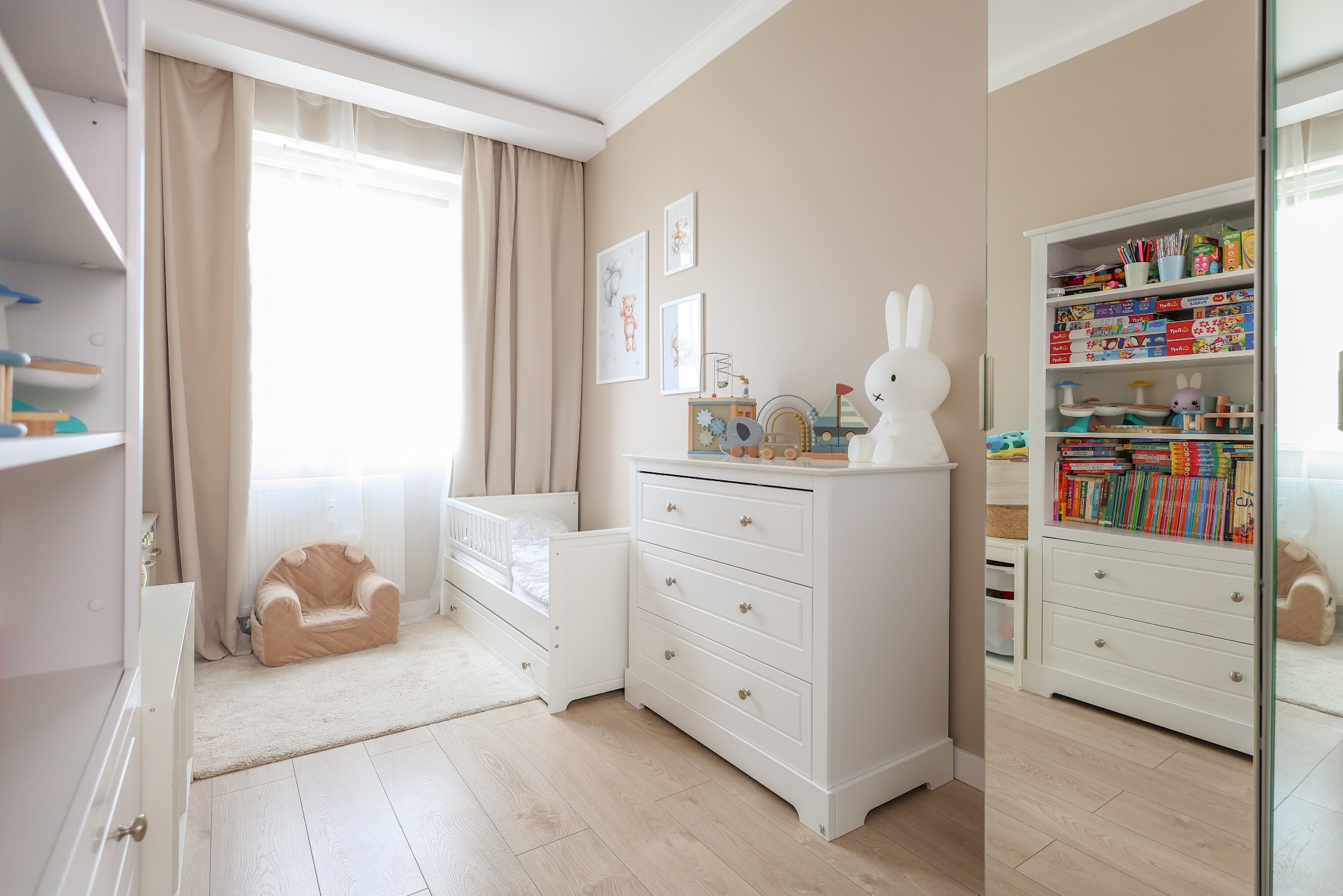 Jasny pokój dziecięcy z białymi meblami: łóżeczko, komoda z zabawkami, regał z książkami i pluszakami. Beżowe ściany, dywan i zasłony tworzą przytulną atmosferę.