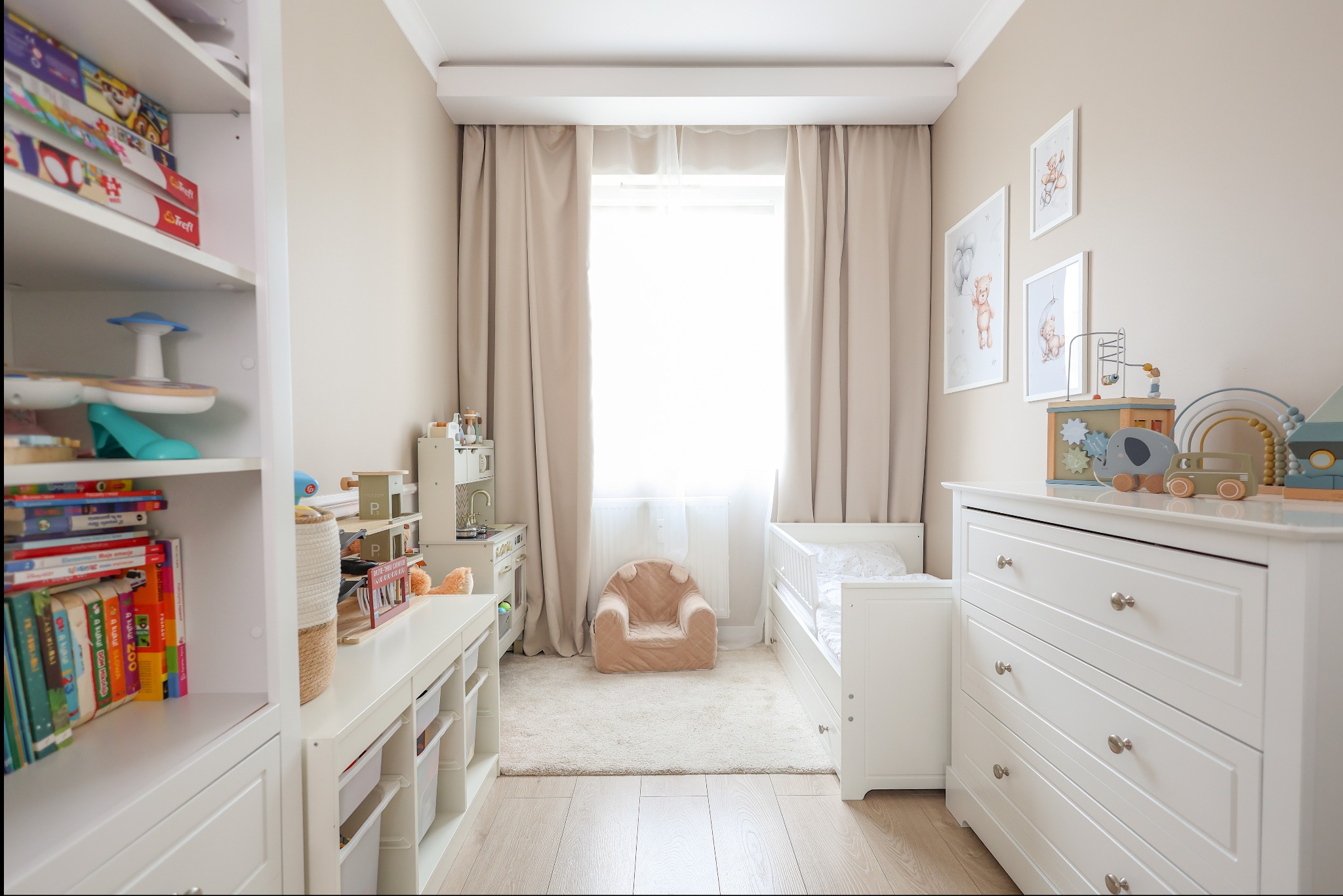 Jasne wnętrze pokoju dziecięcego z białymi meblami, drewnianą podłogą i beżowymi ścianami. Widoczne zabawki, łóżeczko i fotel. Przytulna aranżacja.