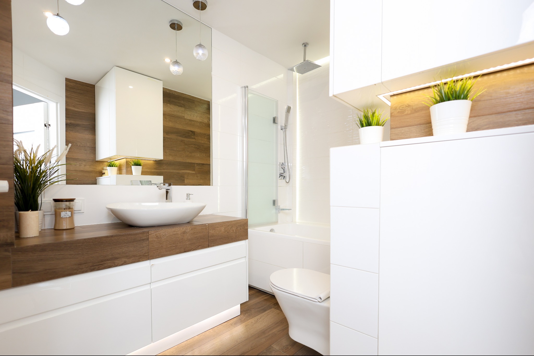 Nowoczesna łazienka z drewnianymi akcentami: białe szafki, owalna umywalka nablatowa, podświetlenie LED, kabina prysznicowa za szklaną ścianką, rośliny doniczkowe.