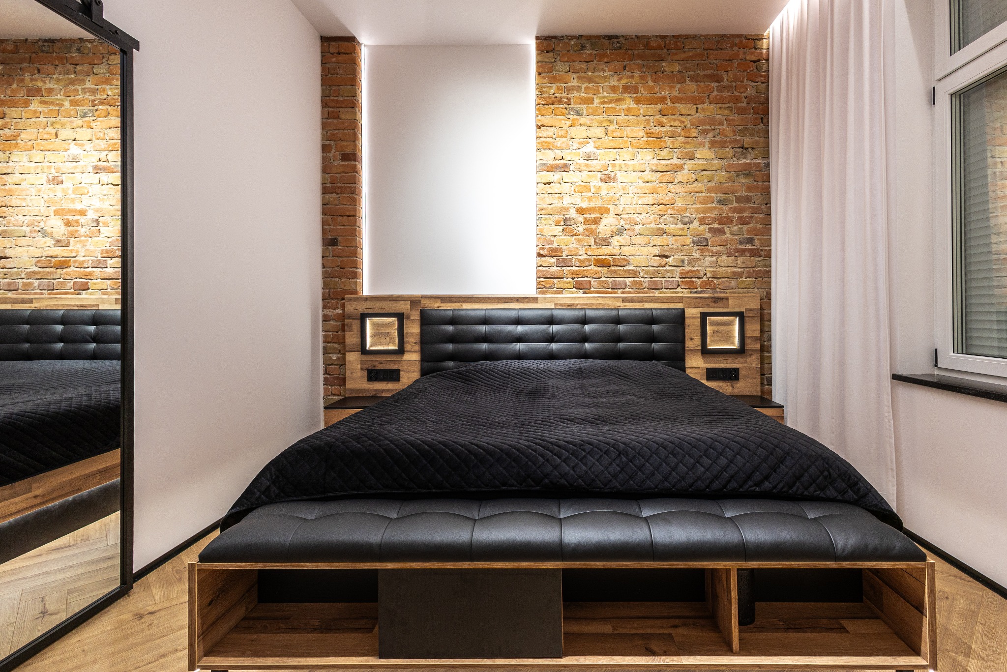 Loftowa sypialnia z ceglaną ścianą, czarnym łóżkiem i lustrem. Widoczne drewniane elementy i oświetlenie LED. Nowoczesny design wnętrza.