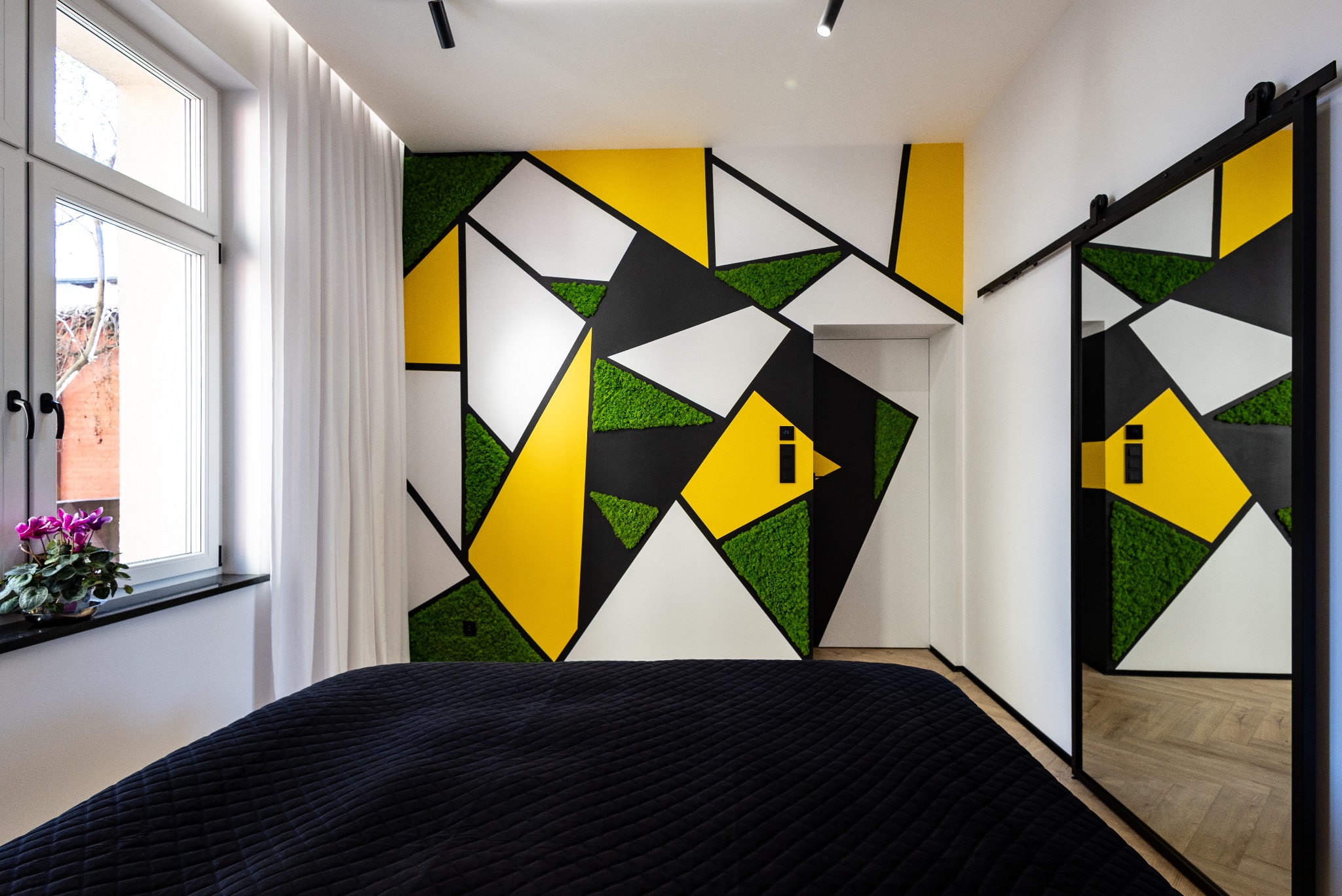 Nowoczesna sypialnia z geometryczną dekoracją ściany z mchu, czerni, bieli i żółci. Przesuwne drzwi lustrzane odbijają wzór. Kwiaty na parapecie, czarna narzuta.