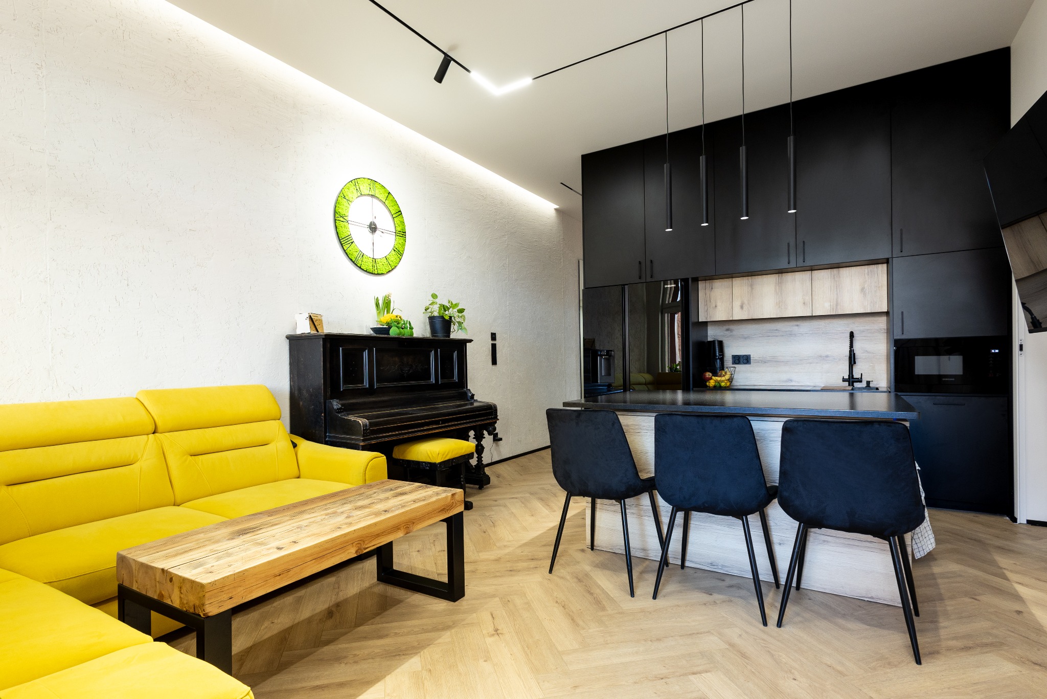 Nowoczesne wnętrze: żółta sofa, drewniany stolik, czarne pianino i kuchnia z wyspą na parkiecie w jodełkę. Zegar na ścianie dodaje koloru.