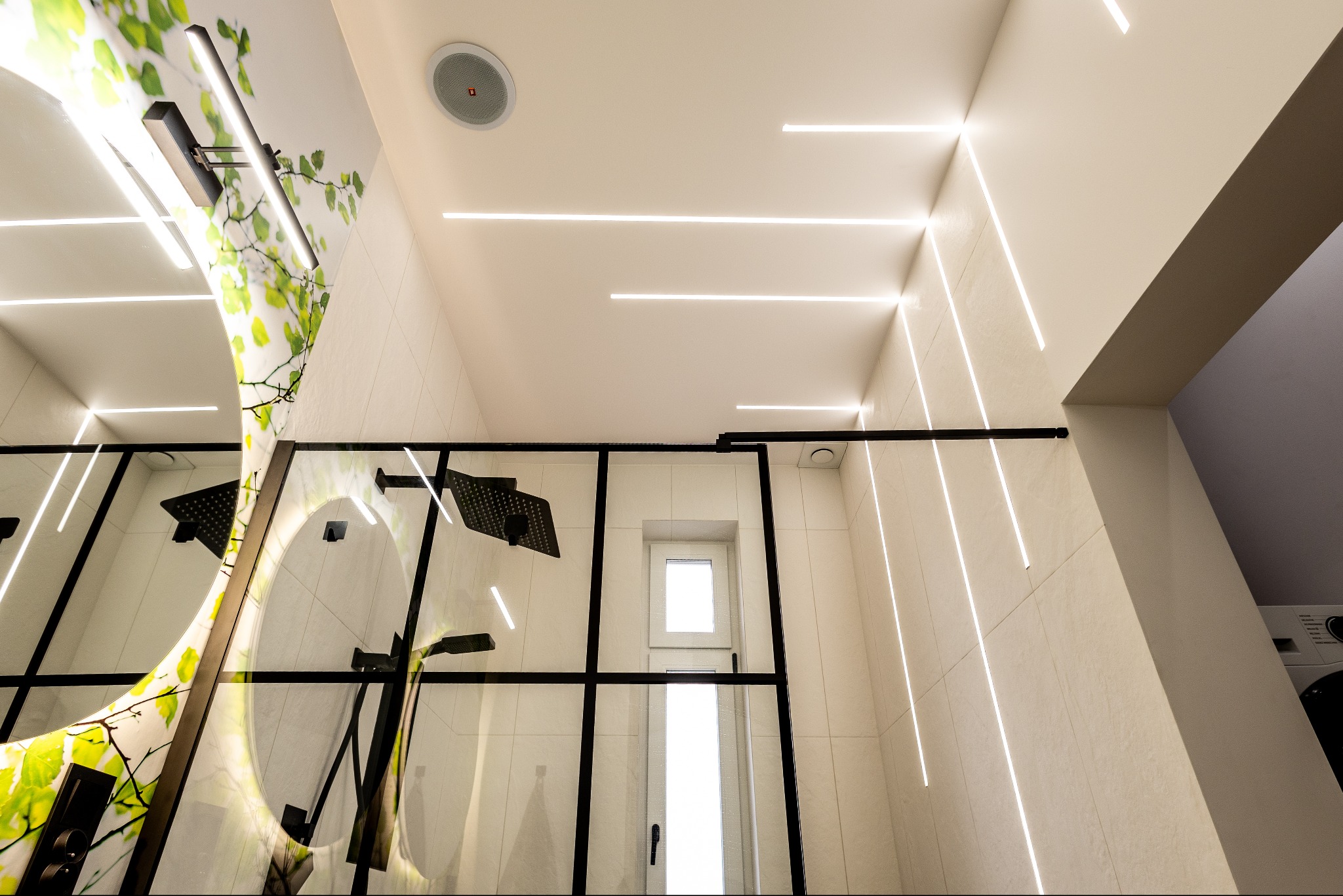 Nowoczesna łazienka z kabiną typu walk-in, podświetleniem LED w suficie i ścianach, oraz okrągłym lustrem z motywem roślinnym. Widok z dołu.