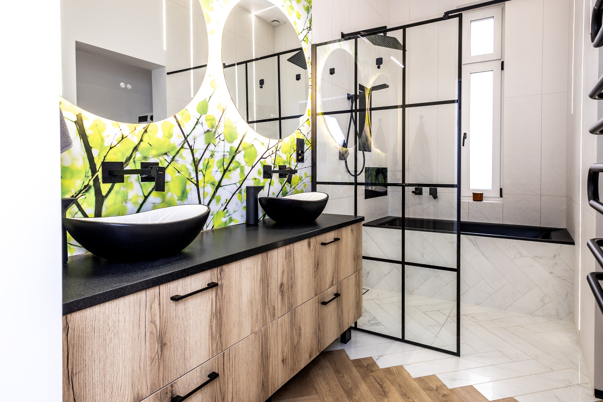Nowoczesna łazienka z drewnianą szafką pod umywalki, okrągłym lustrem z podświetleniem i kabiną prysznicową typu walk-in z czarnymi profilami.