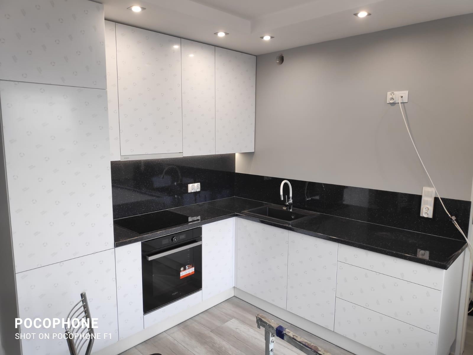Nowoczesna kuchnia z białymi szafkami zdobionymi subtelnym wzorem, czarnym blatem i wbudowanym piekarnikiem. Widoczne gniazdka elektryczne i chromowana bateria.