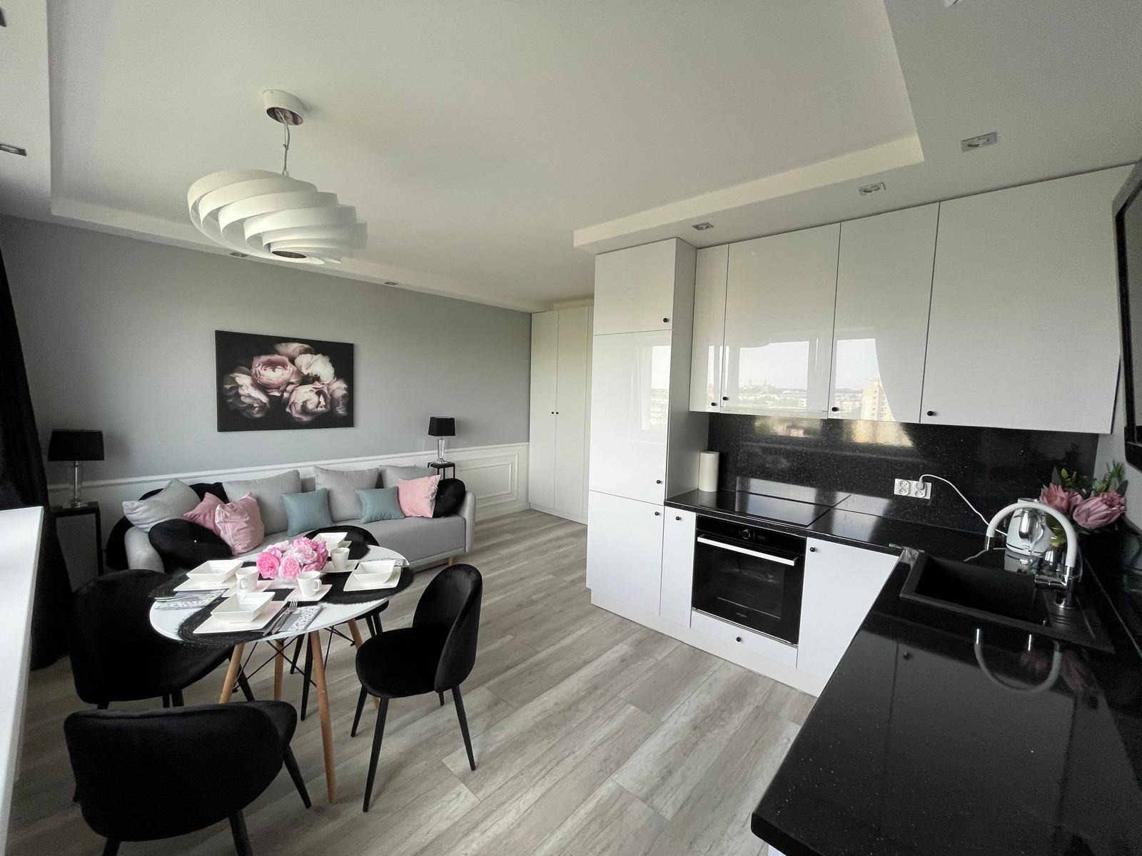 Nowoczesne wnętrze: szara ściana z obrazem kwiatów, sofa z poduszkami, okrągły stół z czarnymi krzesłami i biała kuchnia z czarnym blatem.