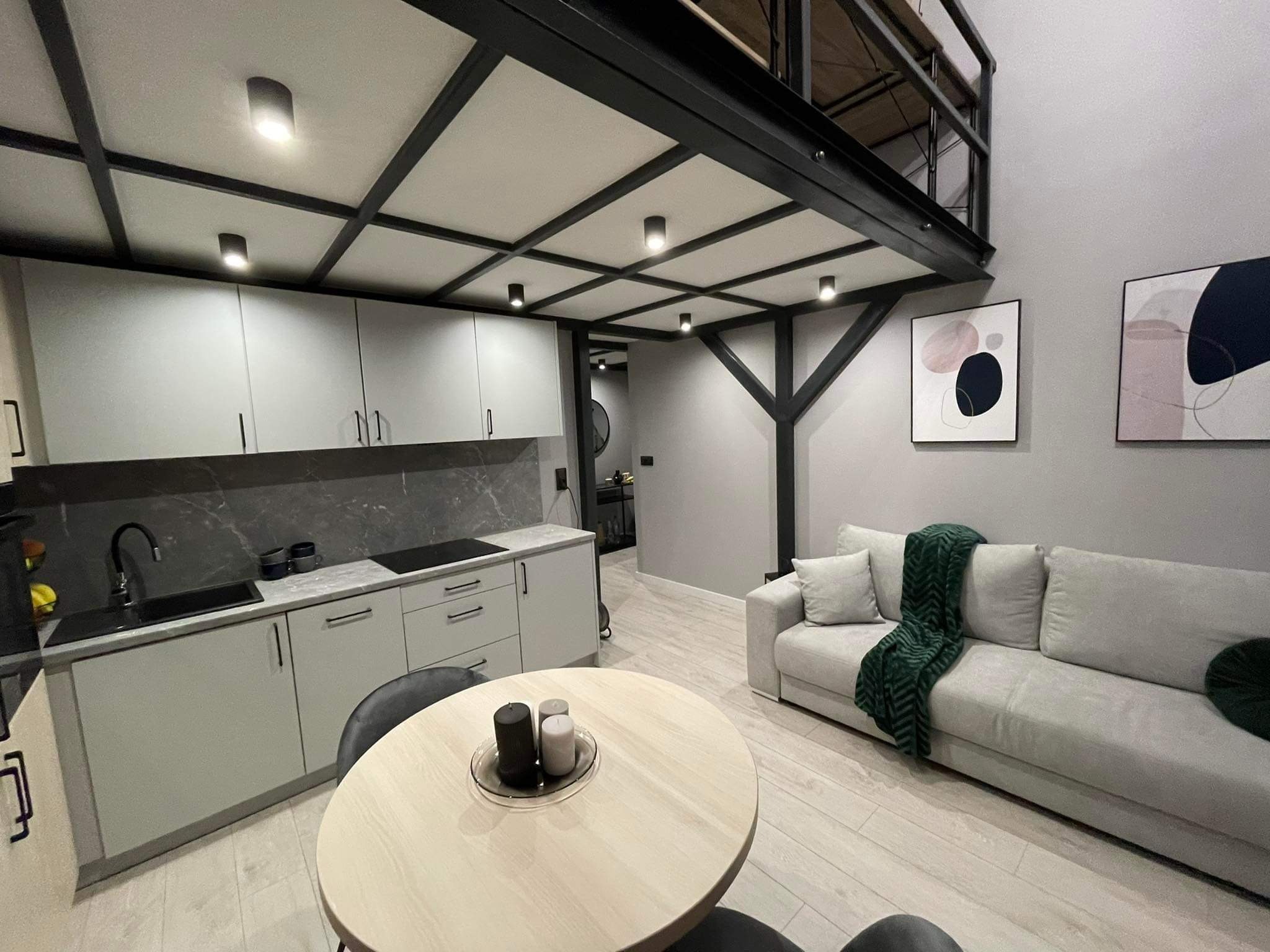 Nowoczesne wnętrze z antresolą: szara kuchnia z marmurowym blatem, okrągły stół z dekoracjami, sofa z zielonym pledem. Czarno-biała, geometryczna dekoracja na ścianie.