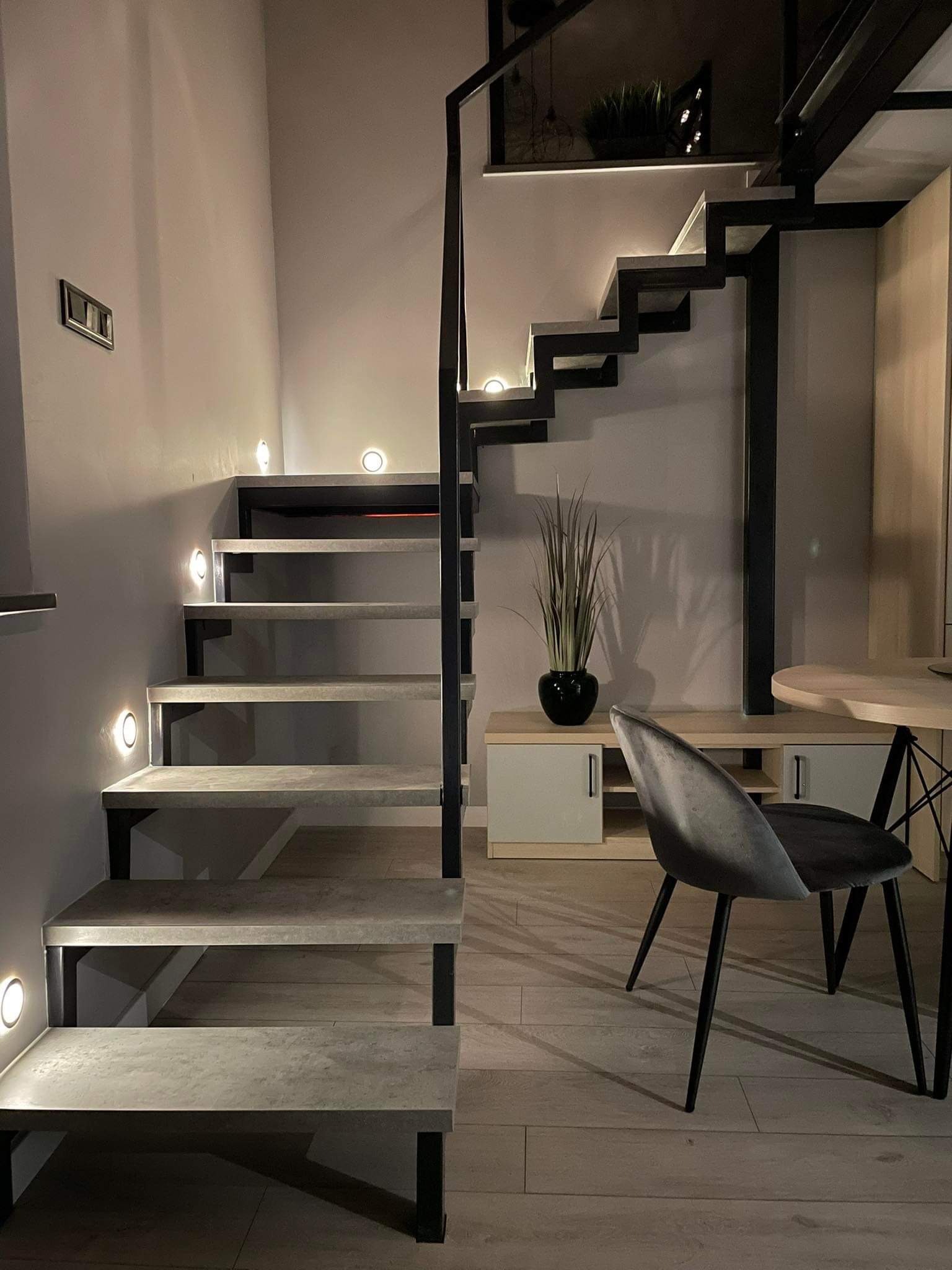 Nowoczesne schody z betonu architektonicznego, podświetlone punktowymi lampami, z czarną metalową konstrukcją, w jasnym wnętrzu z drewnianą podłogą i minimalistycznym wystrojem.