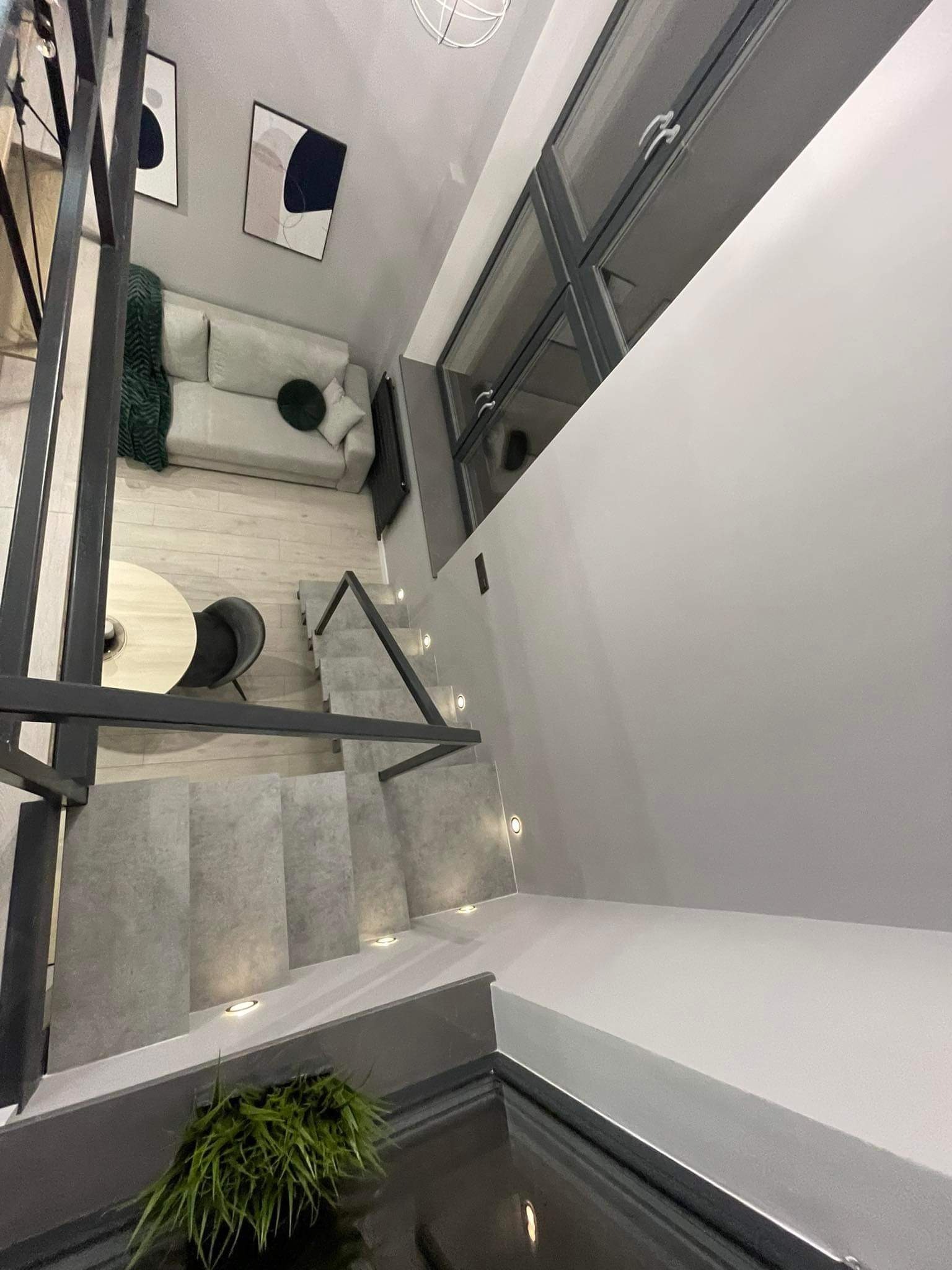 Nowoczesne wnętrze domu z szarymi schodami z podświetleniem LED, widok z góry na salon z sofą i stolikiem kawowym. Minimalistyczny design z obrazami na ścianie.