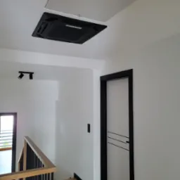 Czarny panel klimatyzacyjny wmontowany w sufit podwieszany w nowoczesnym, minimalistycznym wnętrzu domu, widoczne włączone diody sygnalizacyjne, obok drzwi z czarną ościeżnicą i schody z drewnianą...