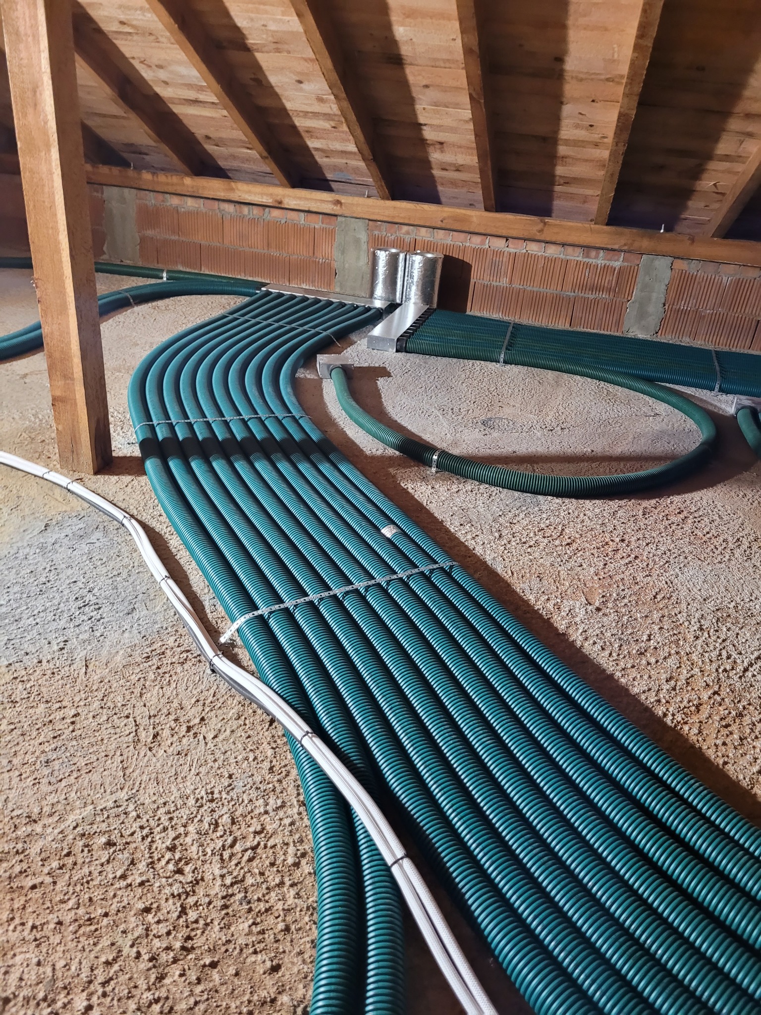 Rozprowadzenie systemu rekuperacji z zielonych, karbowanych rur w przestrzeni poddasza, widoczne elementy mocujące i białe przewody elektryczne.