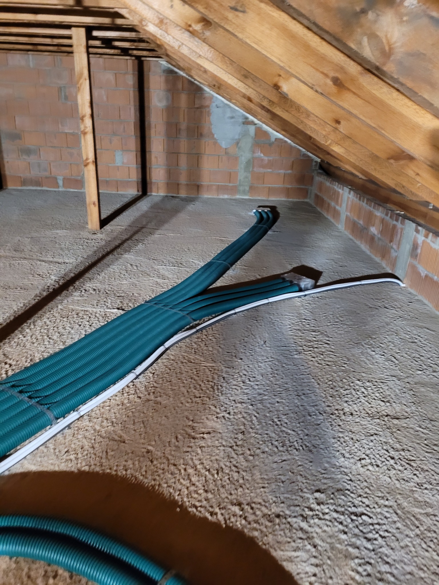 Zielone rury wentylacyjne ułożone na szarej izolacji poddasza, widoczne drewniane krokwie i ceglana ściana.