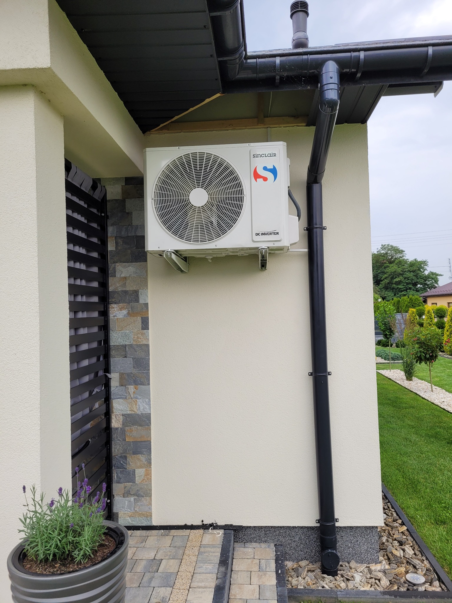 Biała jednostka zewnętrzna klimatyzatora Sinclair zamontowana na ścianie domu z beżowym tynkiem, obok czarnej rynny i dekoracyjnej elewacji z kamienia łupkowego, z widocznym fragmentem ogrodu w tle.