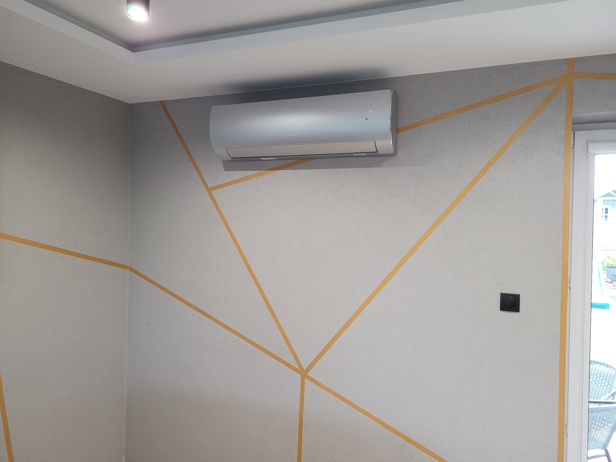 Zamontowana srebrna klimatyzacja ścienna w pokoju z szarymi ścianami ozdobionymi geometrycznym wzorem z żółtych pasków.