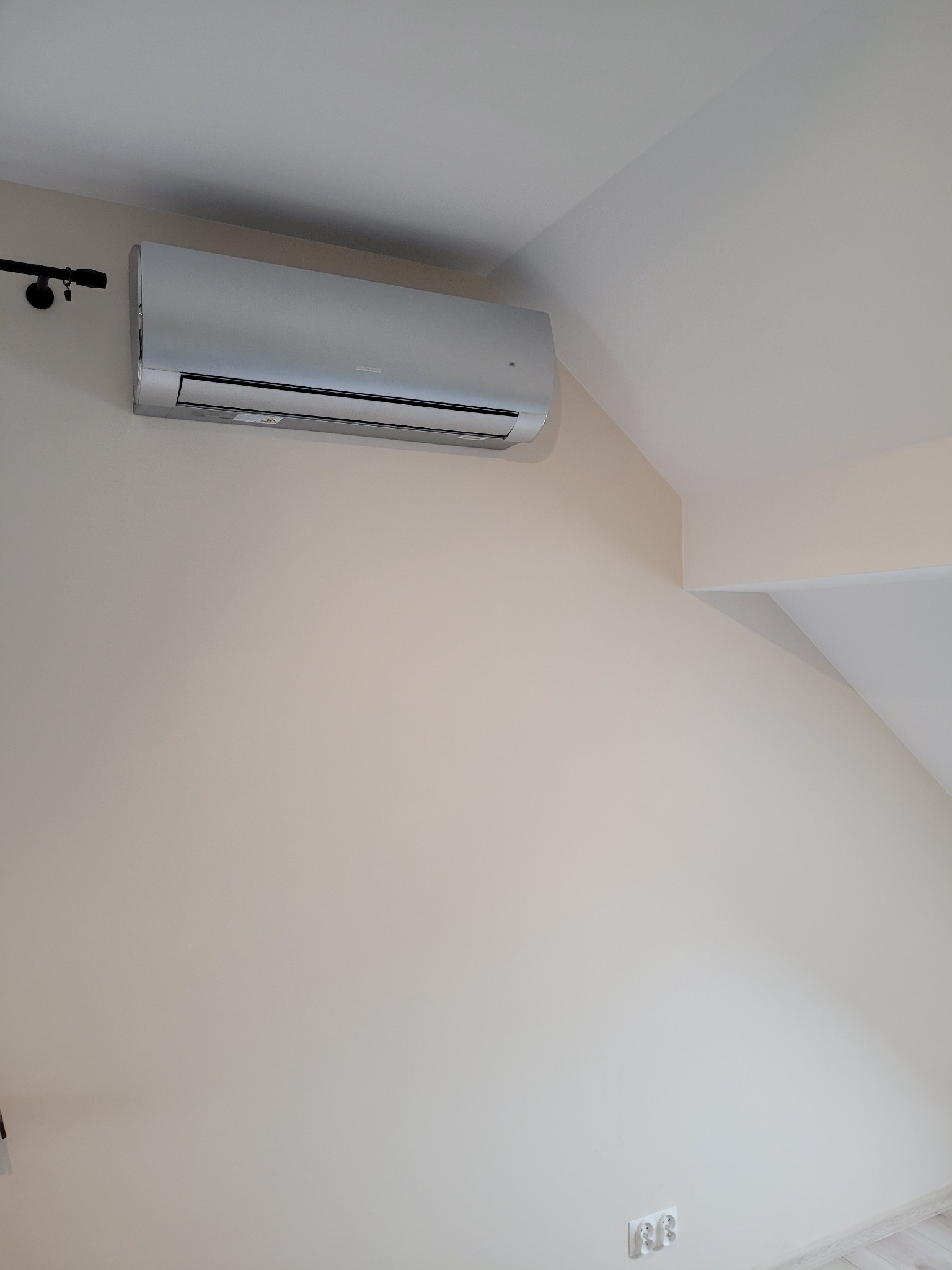 Srebrny klimatyzator ścienny zamontowany w rogu pokoju z beżowymi ścianami i ukośnym sufitem, widoczne gniazdka elektryczne i karnisz.