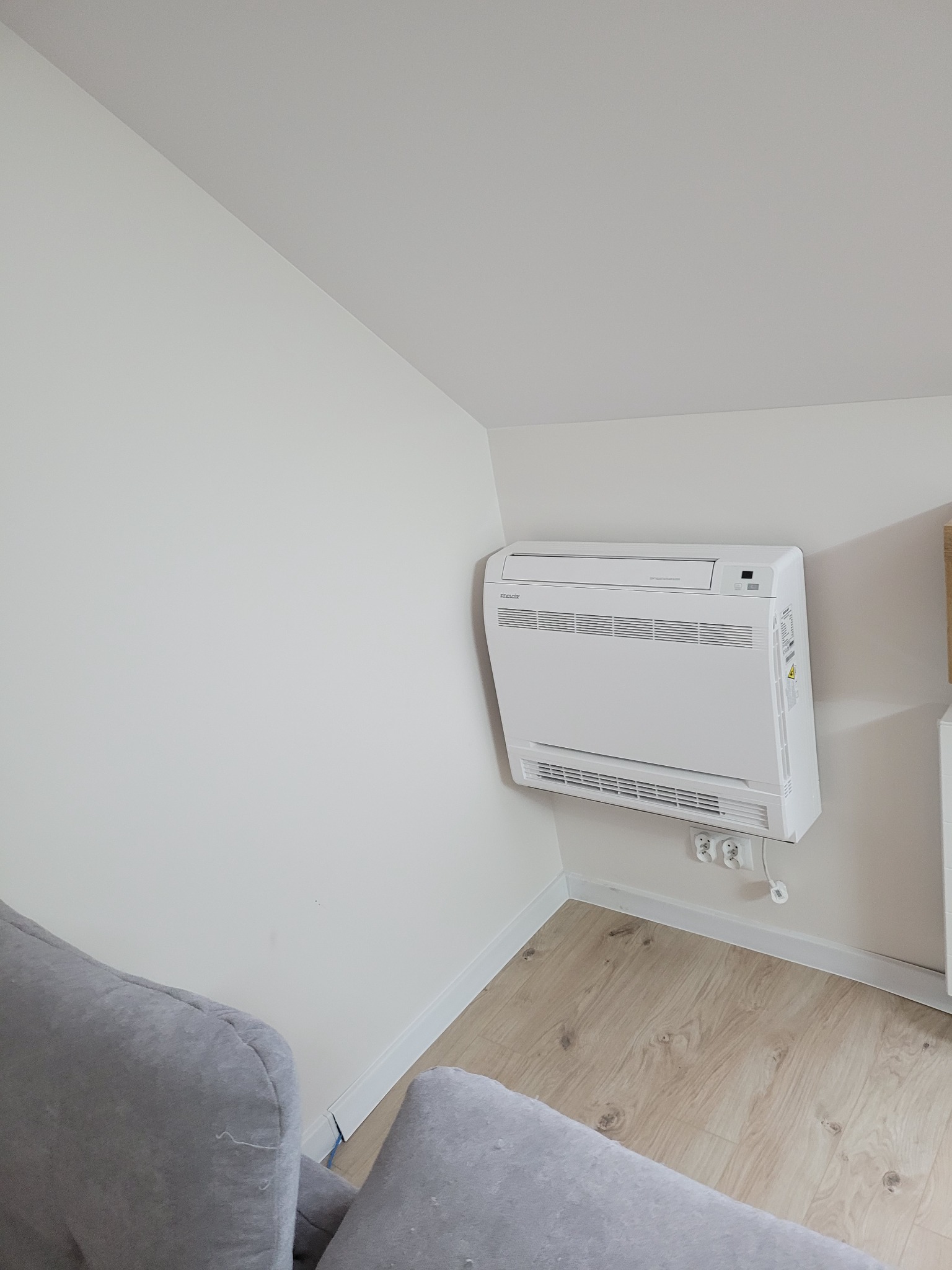 Biały klimatyzator podłogowy zamontowany przy ścianie w jasnym pomieszczeniu, widoczne gniazdka elektryczne i fragment szarego fotela.