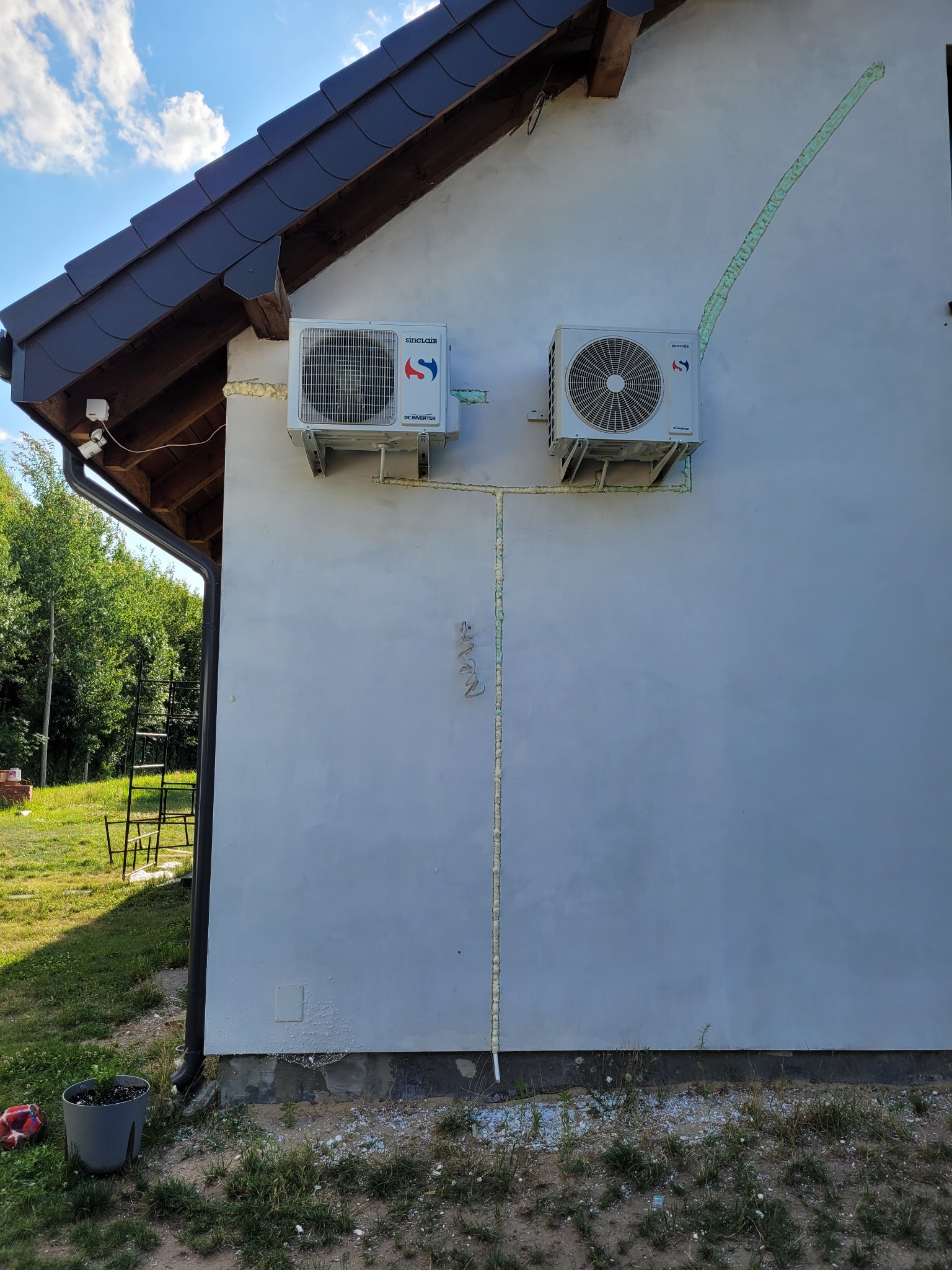 Dwa klimatyzatory Sinclair zamontowane na elewacji domu, rury zabezpieczone pianką montażową, widoczna drabina oparta o ścianę budynku.