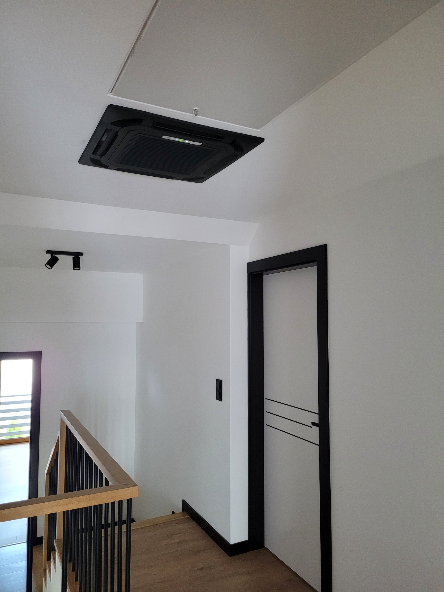 Czarny panel klimatyzacyjny wmontowany w sufit podwieszany w nowoczesnym, minimalistycznym wnętrzu domu, widoczne włączone diody sygnalizacyjne, obok drzwi z czarną ościeżnicą i schody z drewnianą...