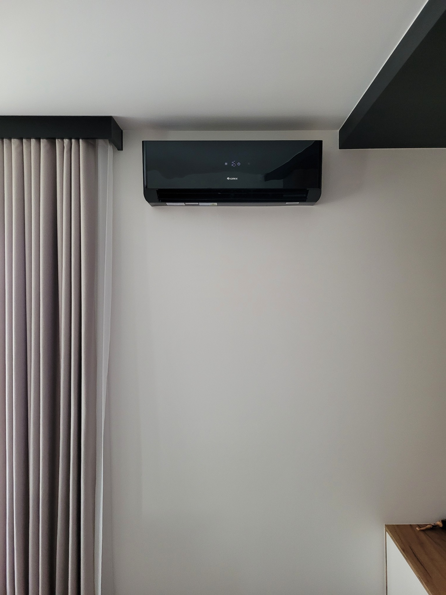 Czarny klimatyzator marki Gree zamontowany na szarej ścianie w pomieszczeniu z zasłoniętymi szarymi zasłonami i czarnym elementem dekoracyjnym pod sufitem.