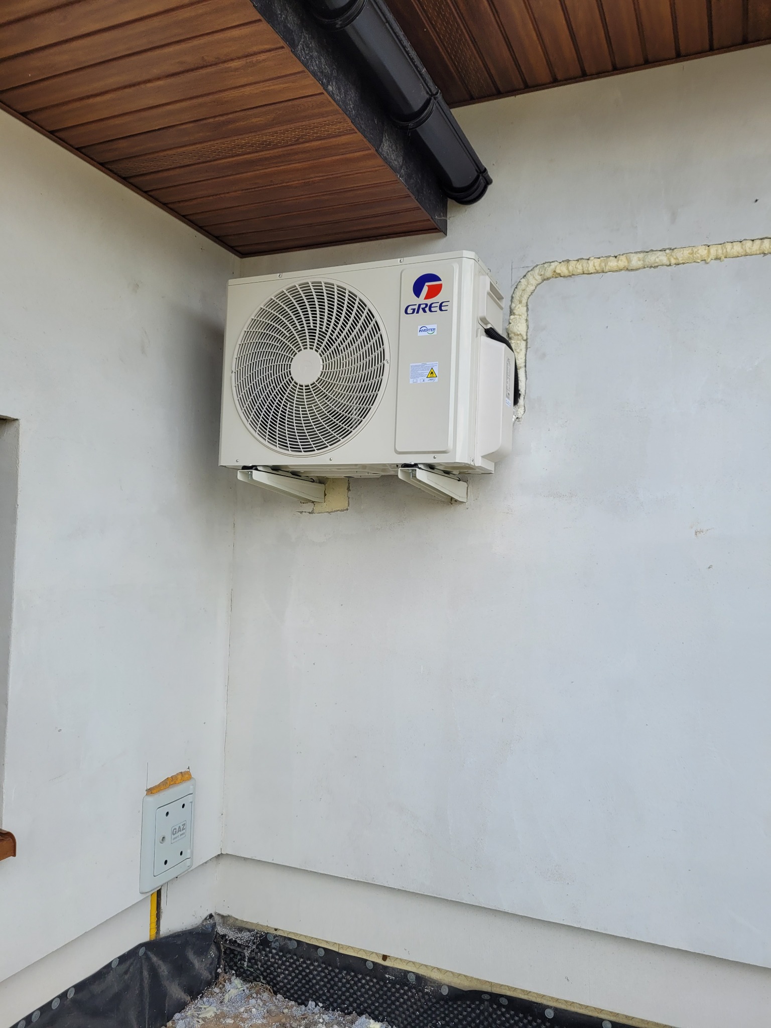 Zewnętrzna jednostka klimatyzacji Gree zamontowana na białej ścianie budynku, z widocznym ociepleniem rur pianką montażową i brązowym podbitką dachu.